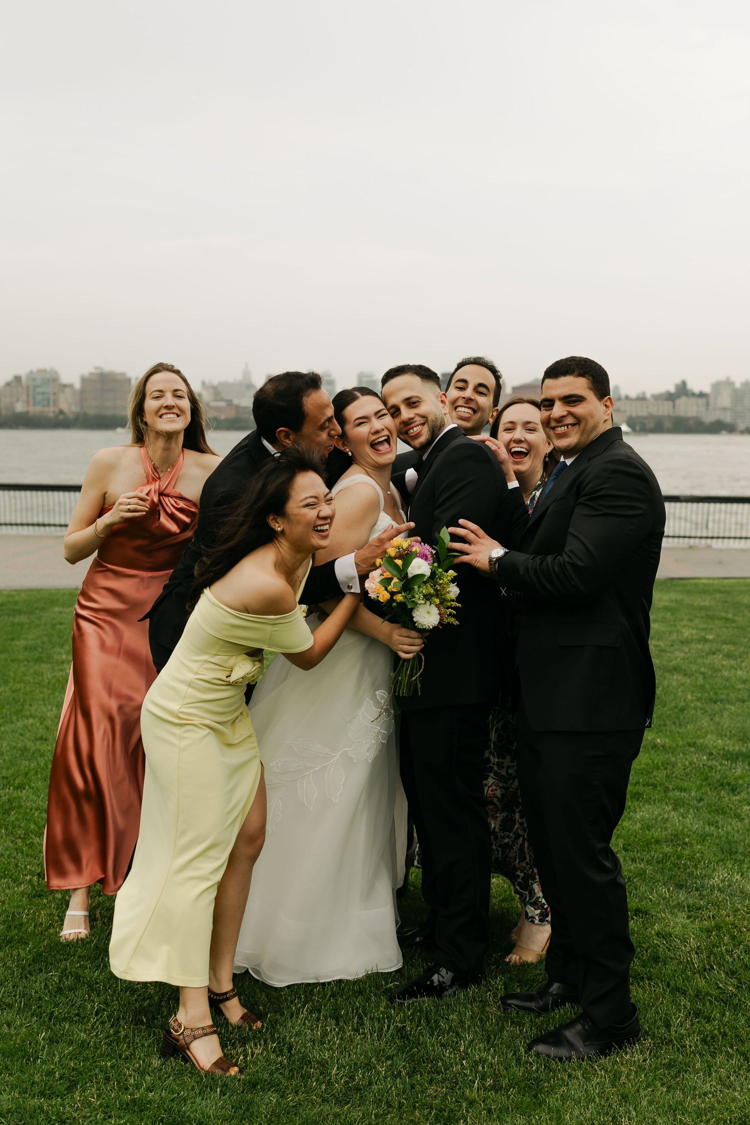 Wedding party celebrates joyfully on grass with cityscape in background during sunset.