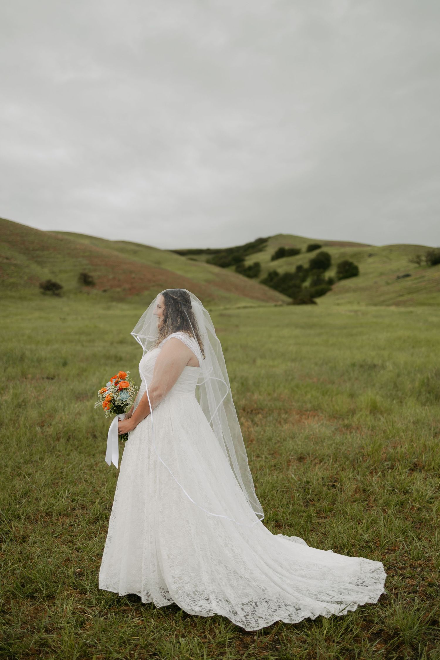 Bride in white lace gown and veil holds orange bouquet in lush green rolling hills under overcast sky.