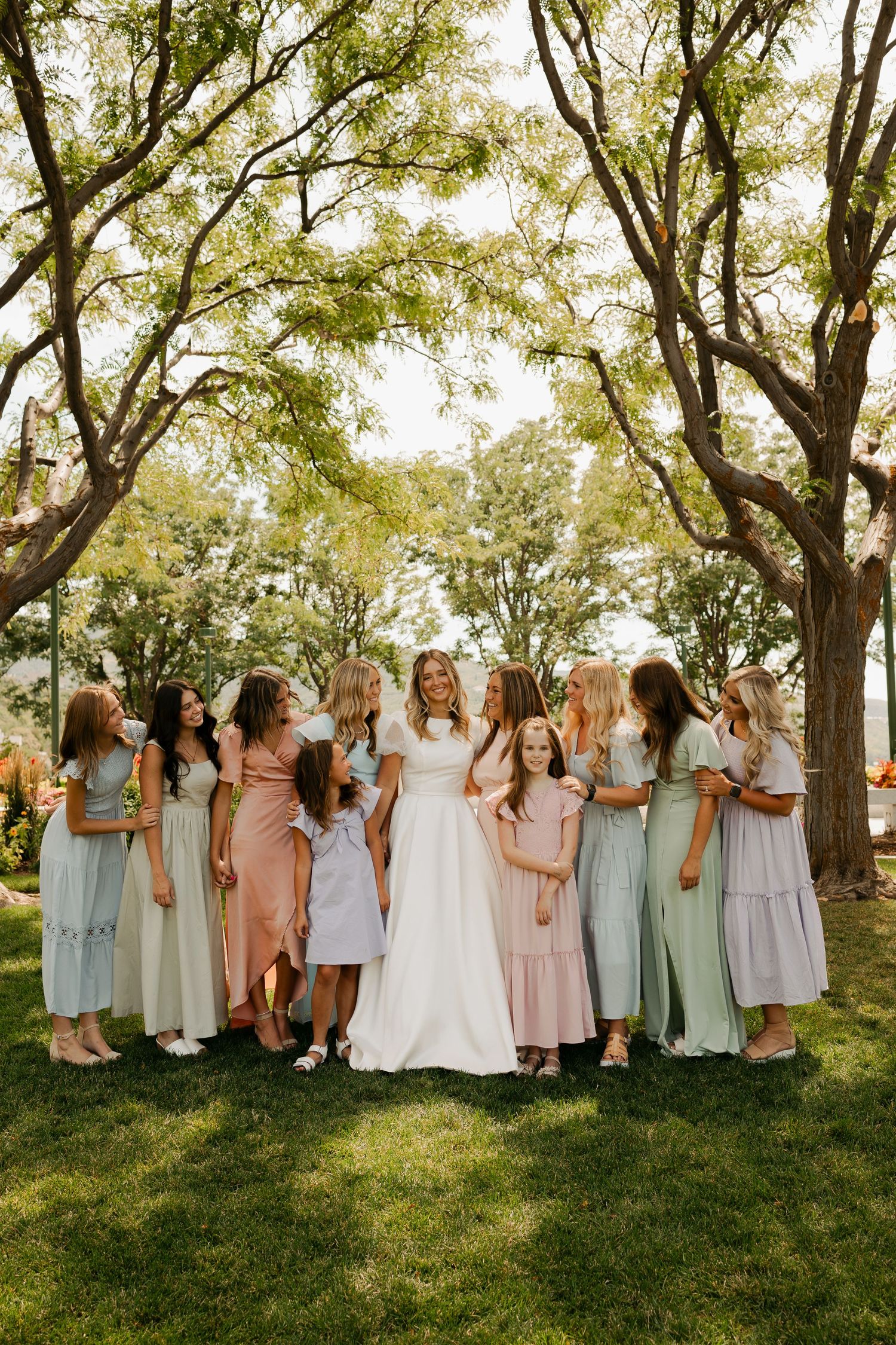 A wedding party in pastel bridesmaid dresses stands together on a grassy hill under large trees.