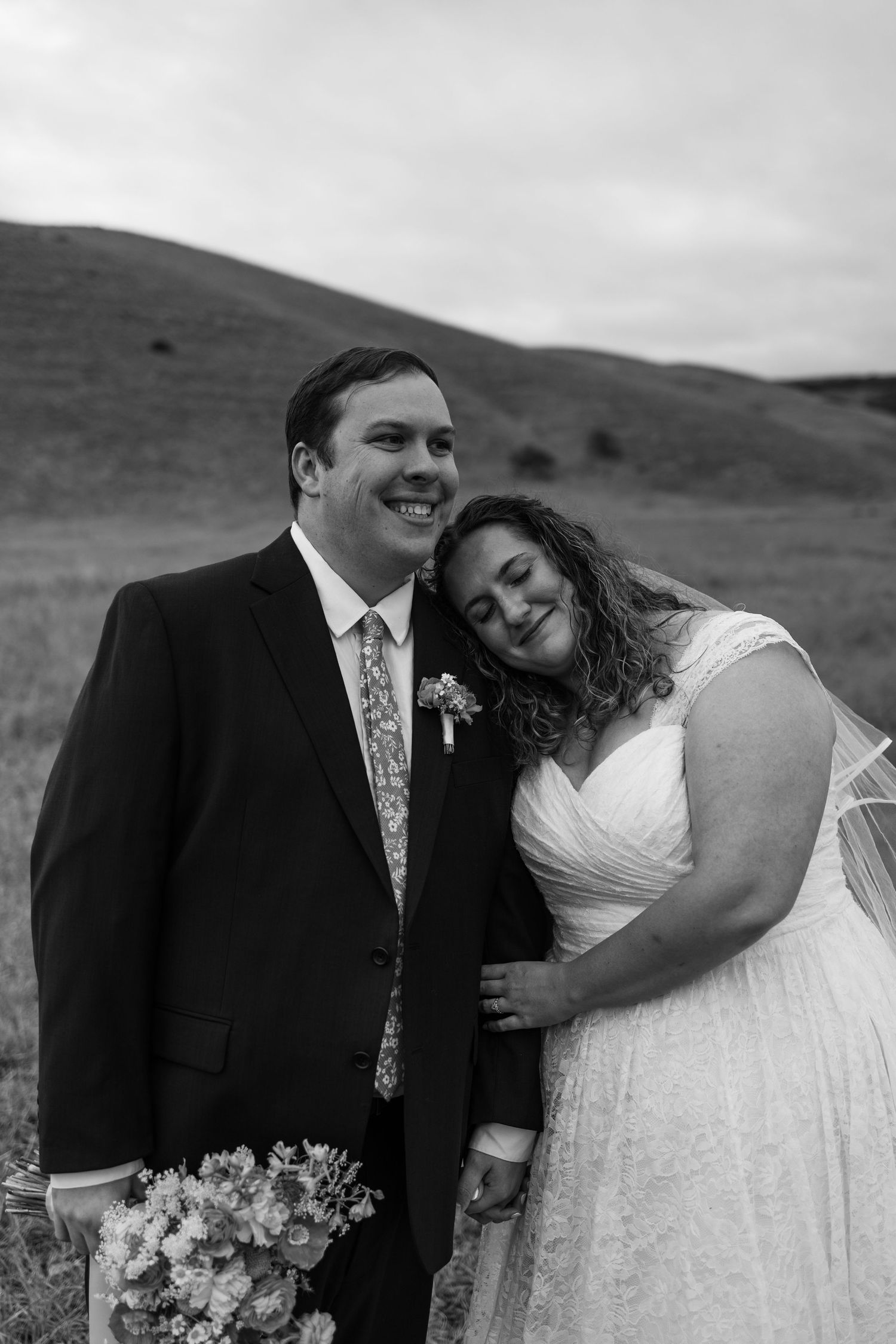 Black and white wedding photo of smiling groom in suit and bride in white dress leaning on his shoulder in a field.