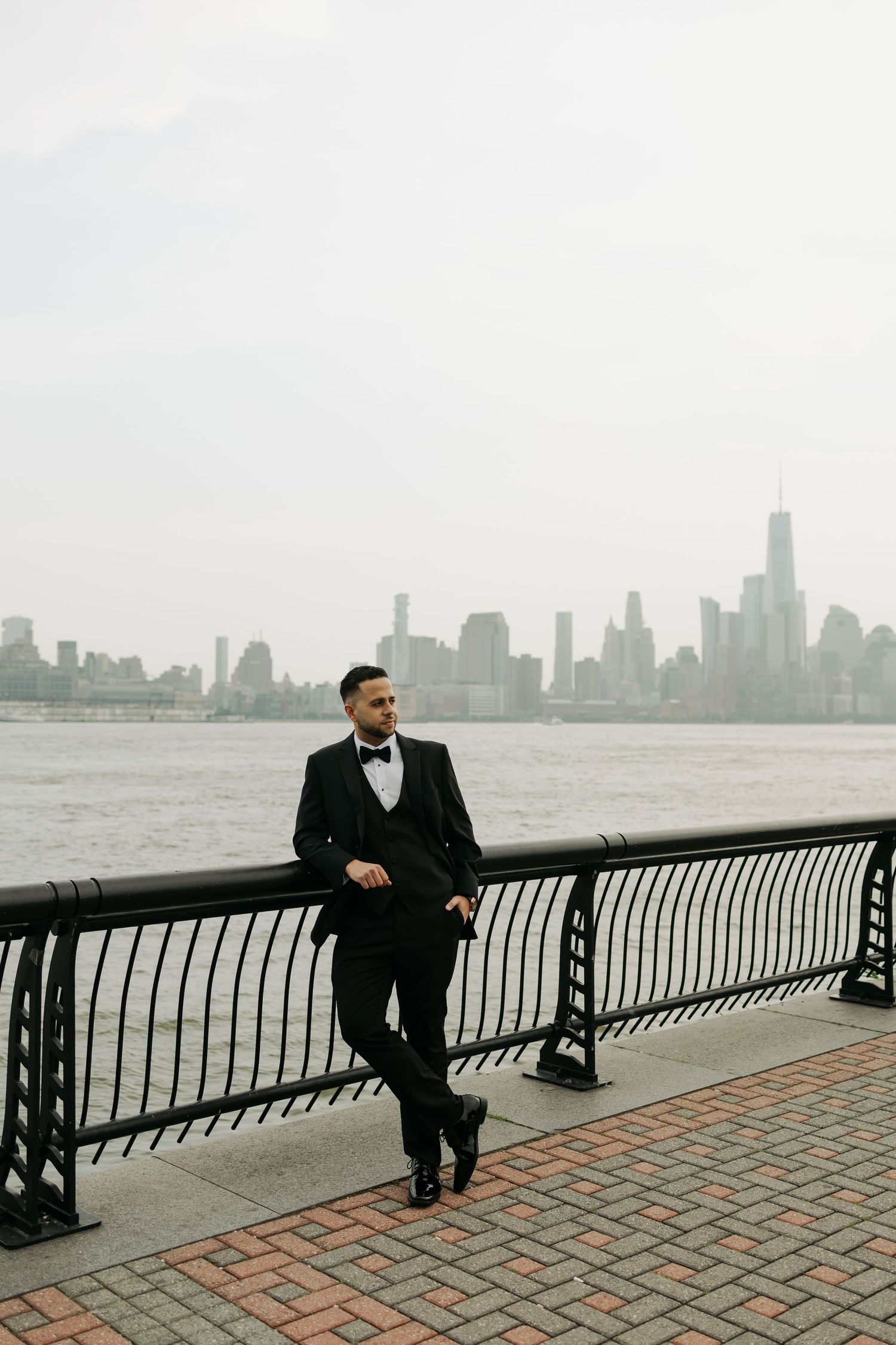 Person in formal attire rests against railing on waterfront path with foggy city skyline in background.