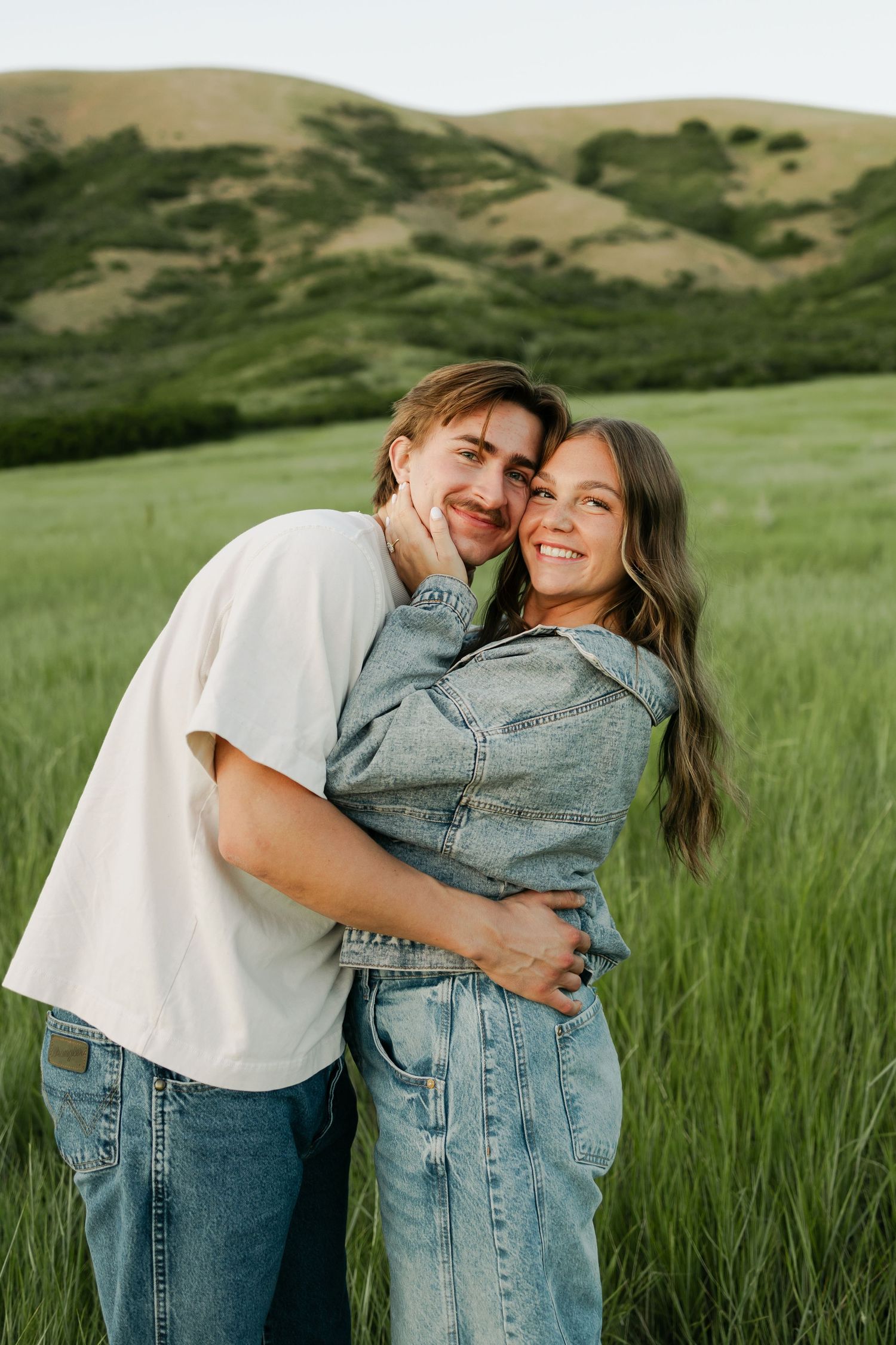 Casual couple embracing and laughing in green field with rolling hills backdrop.