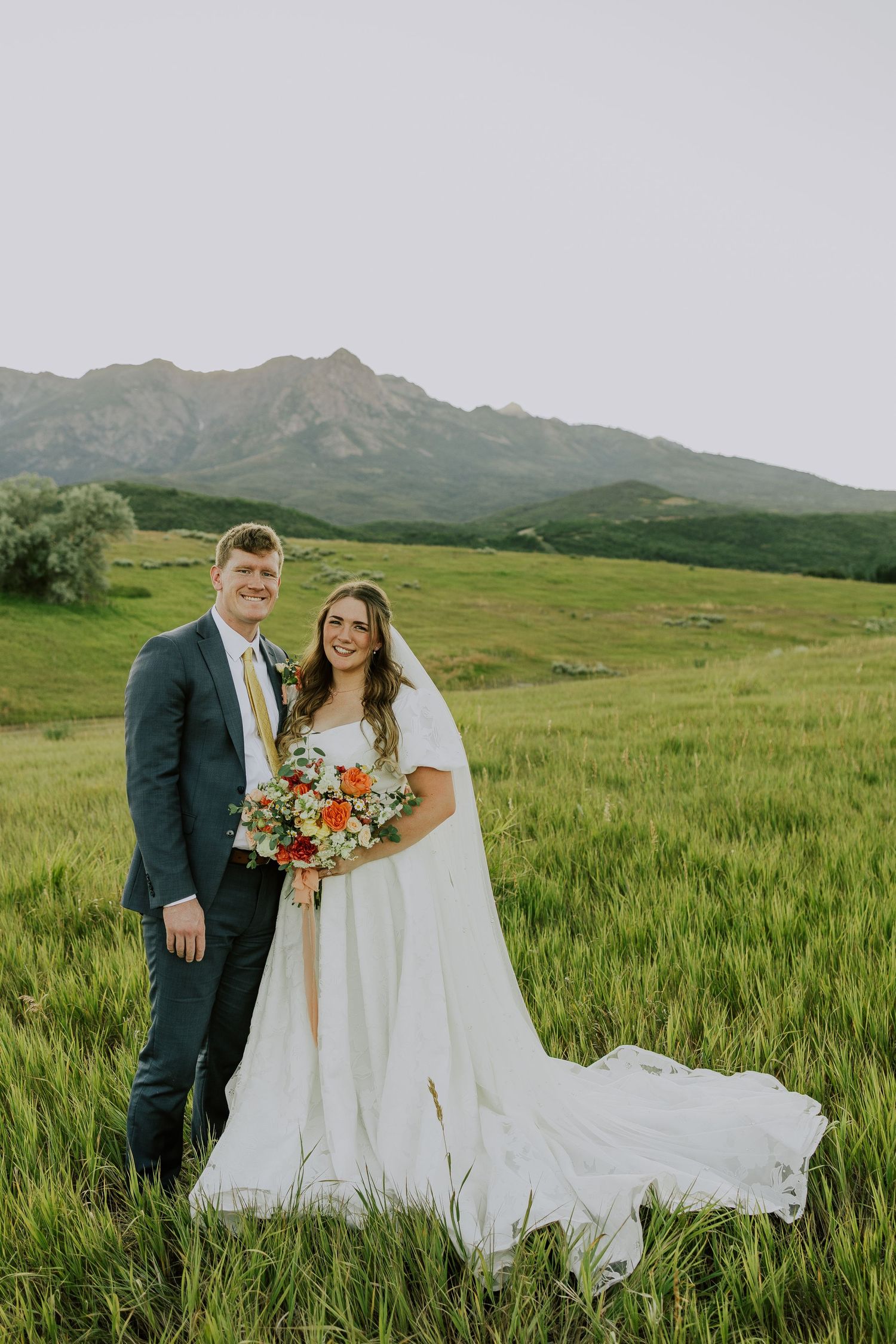 A couple in wedding attire pose together in a grassy meadow with majestic mountain peaks in the background.