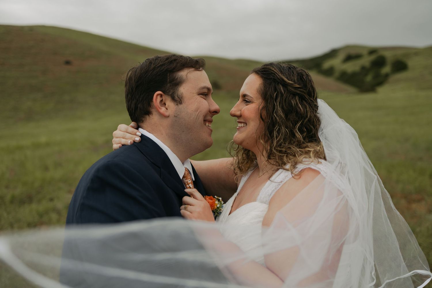 A bride and groom share a joyful moment in a lush green hillside, her veil flowing in the wind.