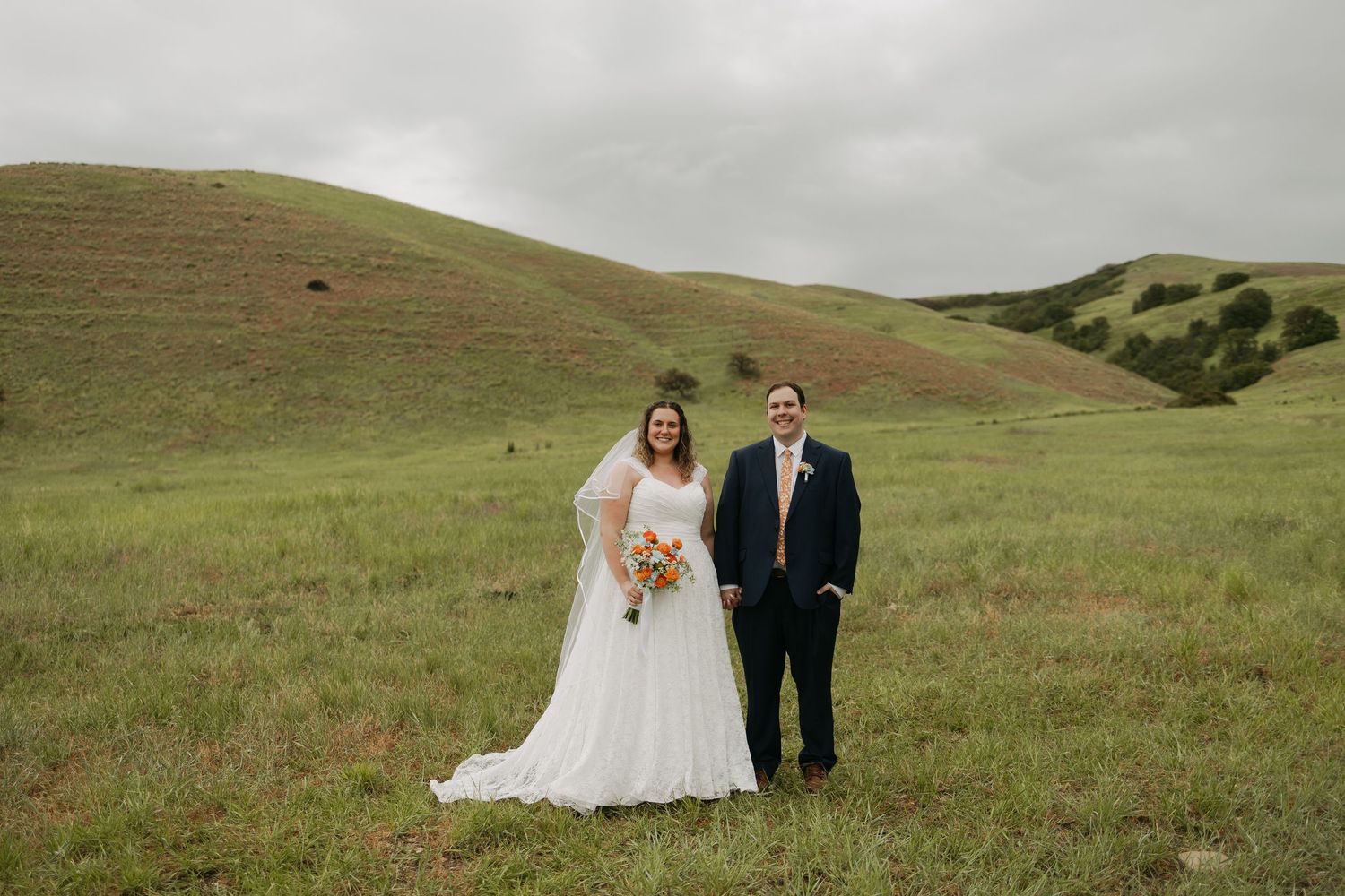 Bride and groom holding bouquet stand in lush green rolling hills on their wedding day.