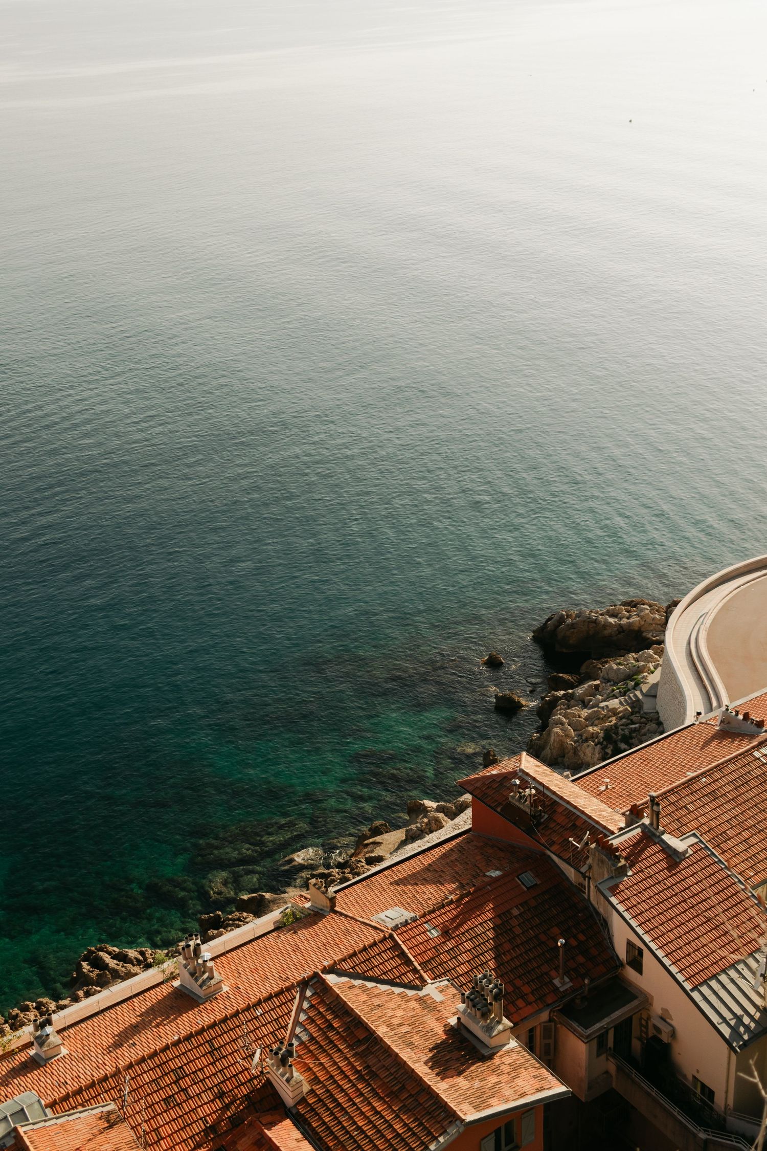 Aerial view of terracotta rooftops overlooking turquoise Mediterranean sea waters with rocky coastline in a coastal village.