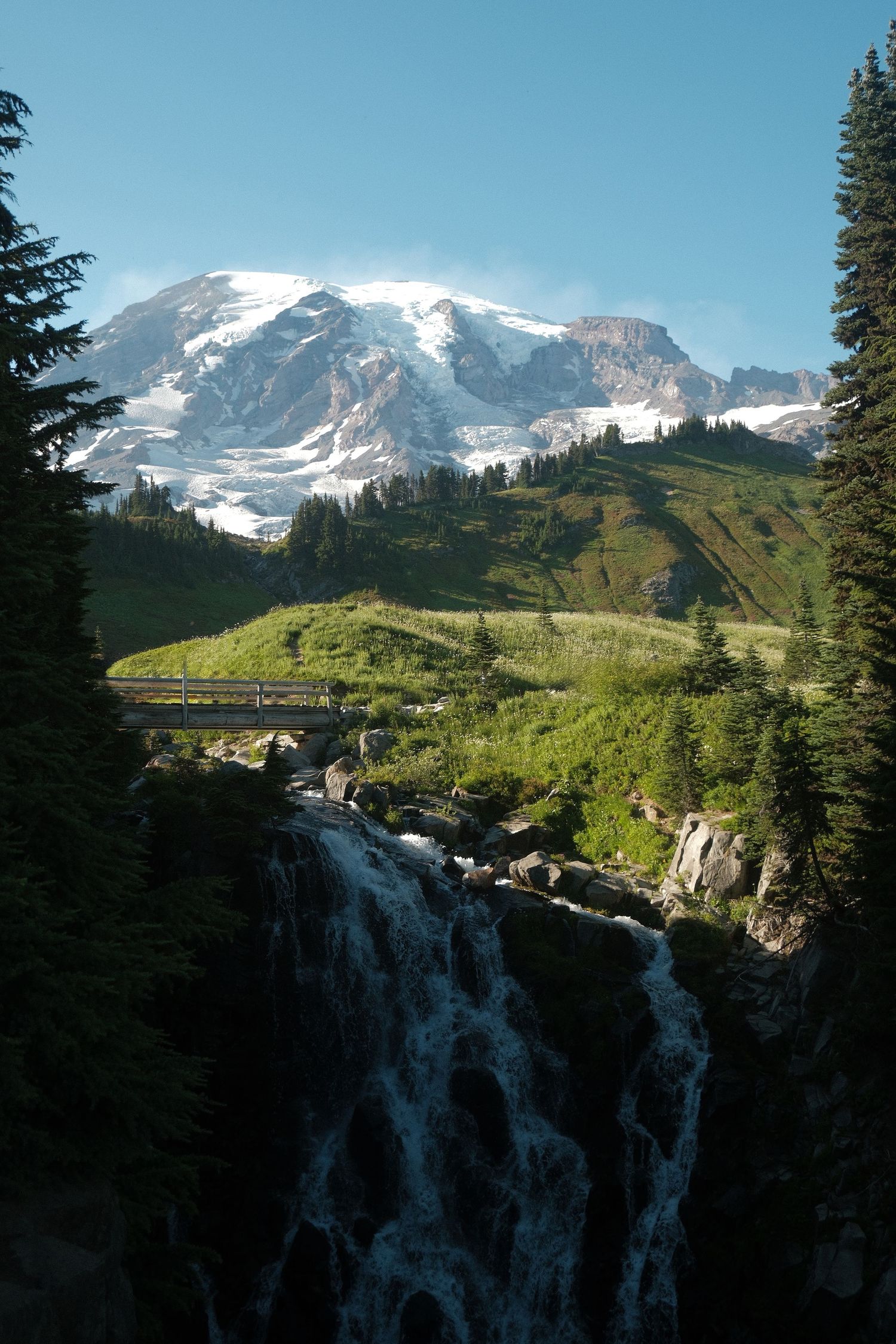 Snow-capped Mount Rainier towers over a lush green valley with a waterfall, bridge, and evergreen trees under a clear blue sky.
