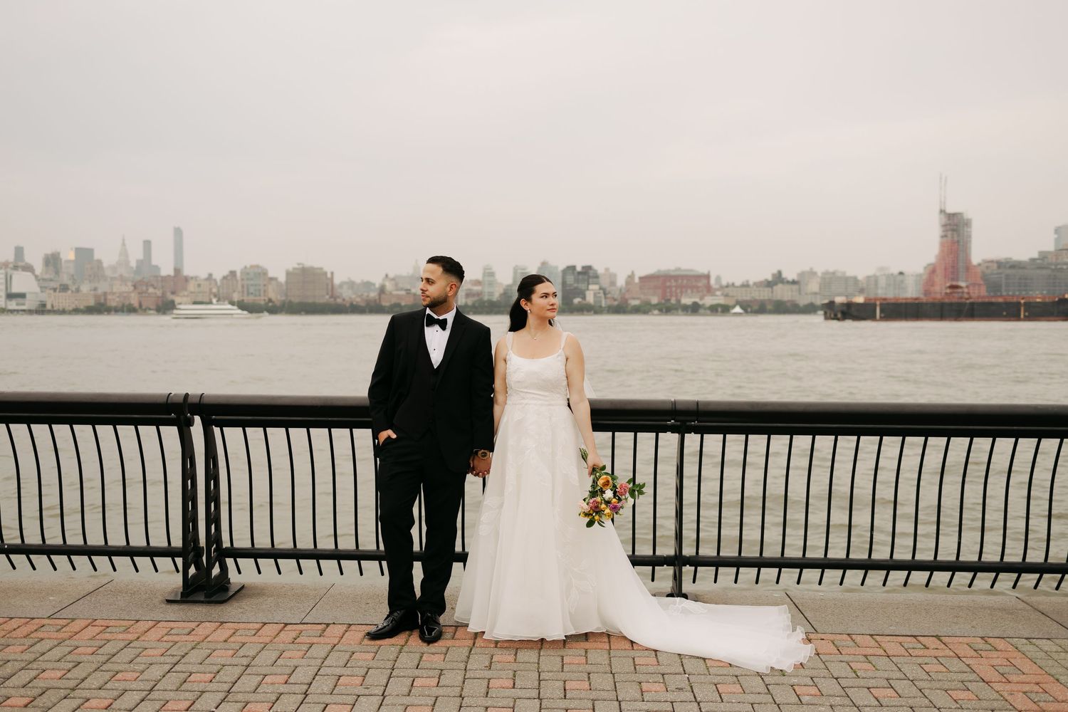 A couple poses together on a waterfront promenade with the city skyline visible across the harbor on an overcast day.