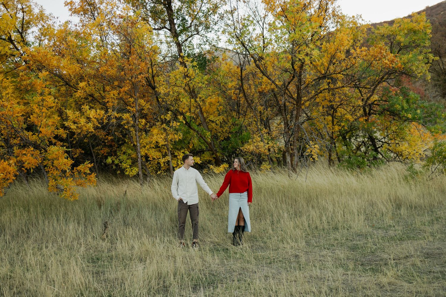 Couple walking hand in hand through fall meadow with autumn trees in background.