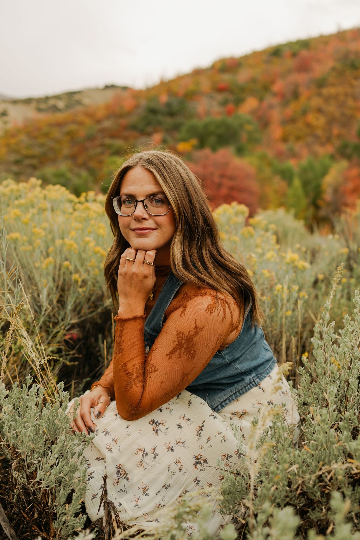 Person in autumn sweater rests head on hand among wildflowers with fall foliage behind.