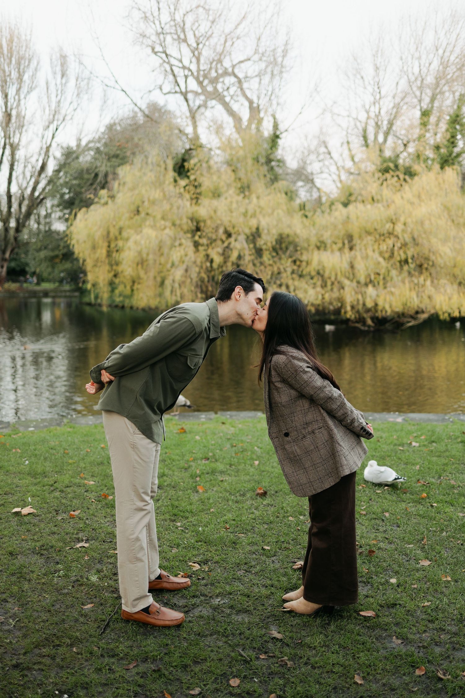 Couple sharing a kiss by a tranquil pond with golden willow trees in the background.