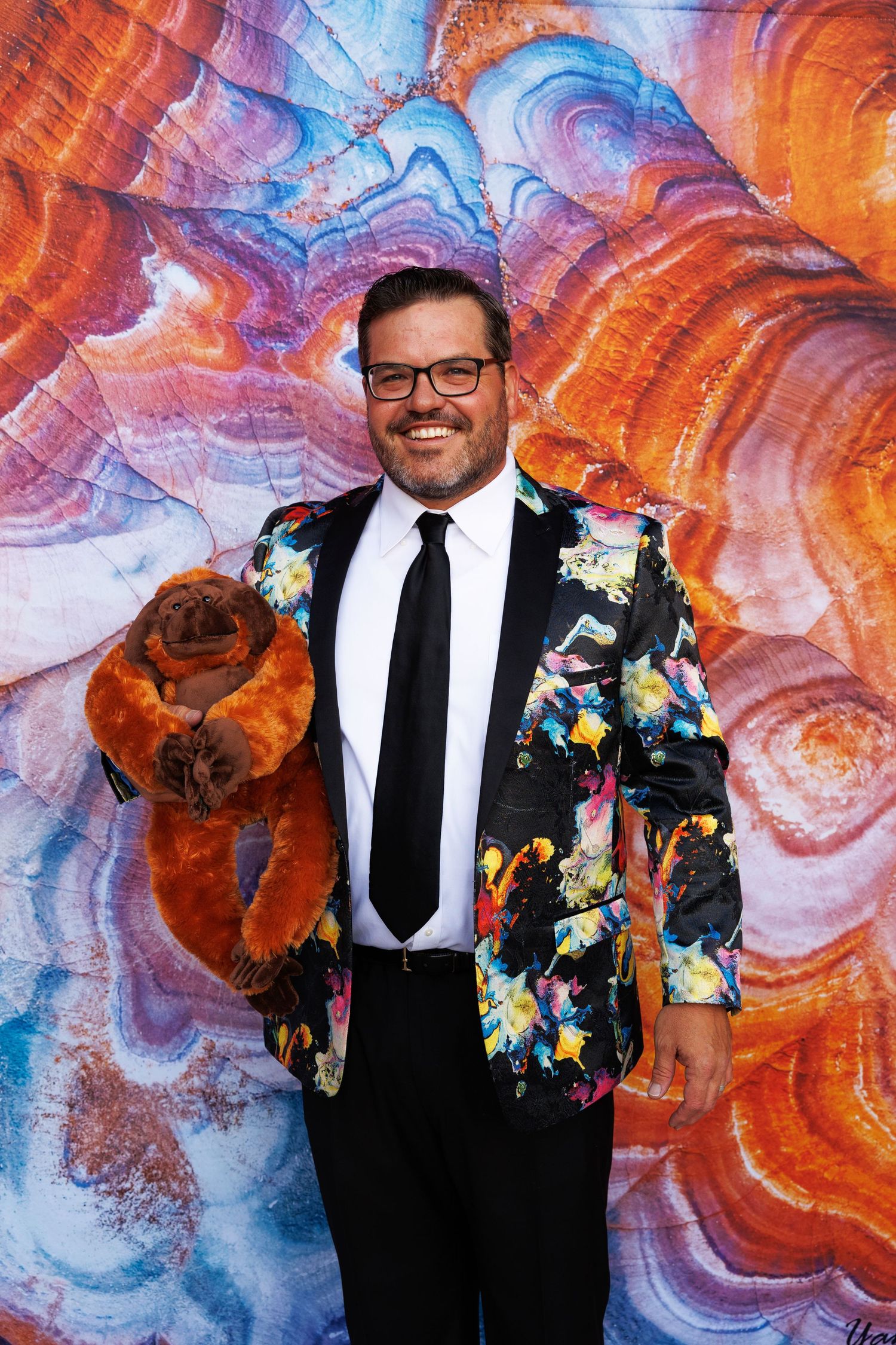 Man in colorful floral blazer holding a stuffed animal, smiling in front of a vibrant marbled background.