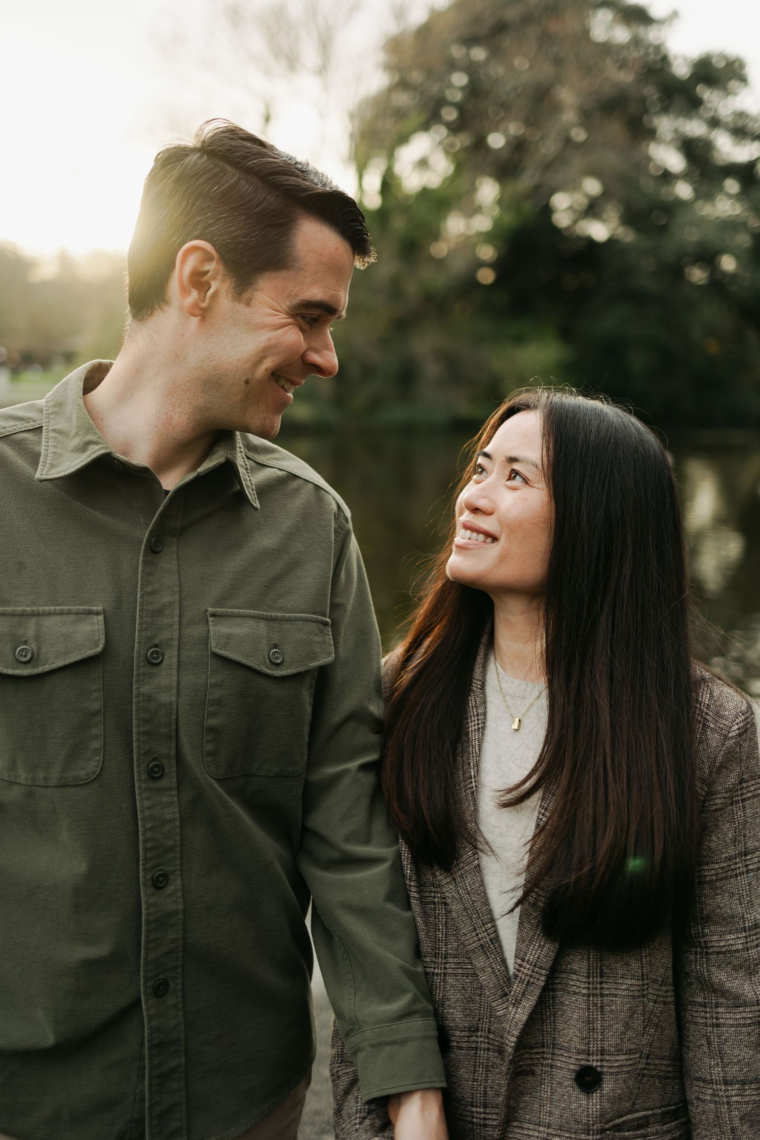 Happy interracial couple smiling at each other in a sunlit park, man in green shirt and woman in plaid blazer.