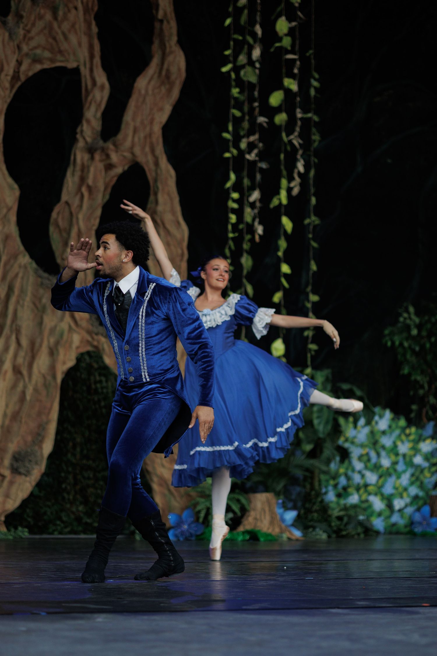 Ballet dancers perform an elegant duet in matching blue costumes against a forest-themed stage backdrop.