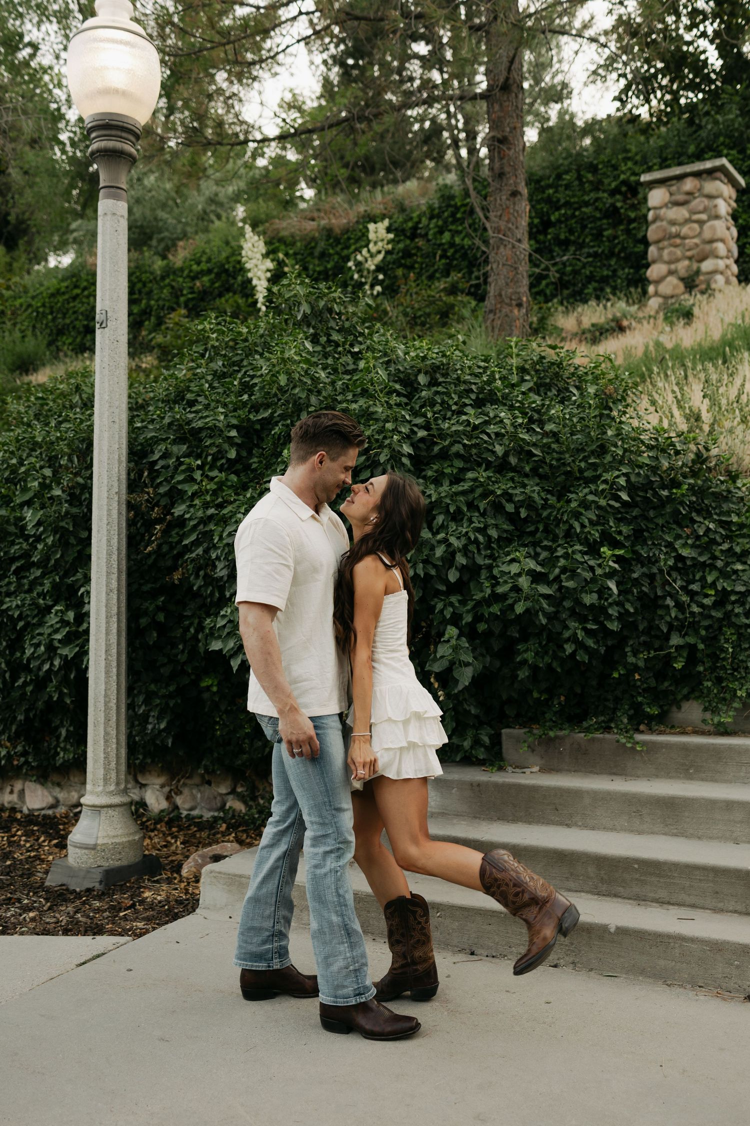 A romantic couple shares a tender moment on concrete steps next to a vintage-style lamppost and green foliage.