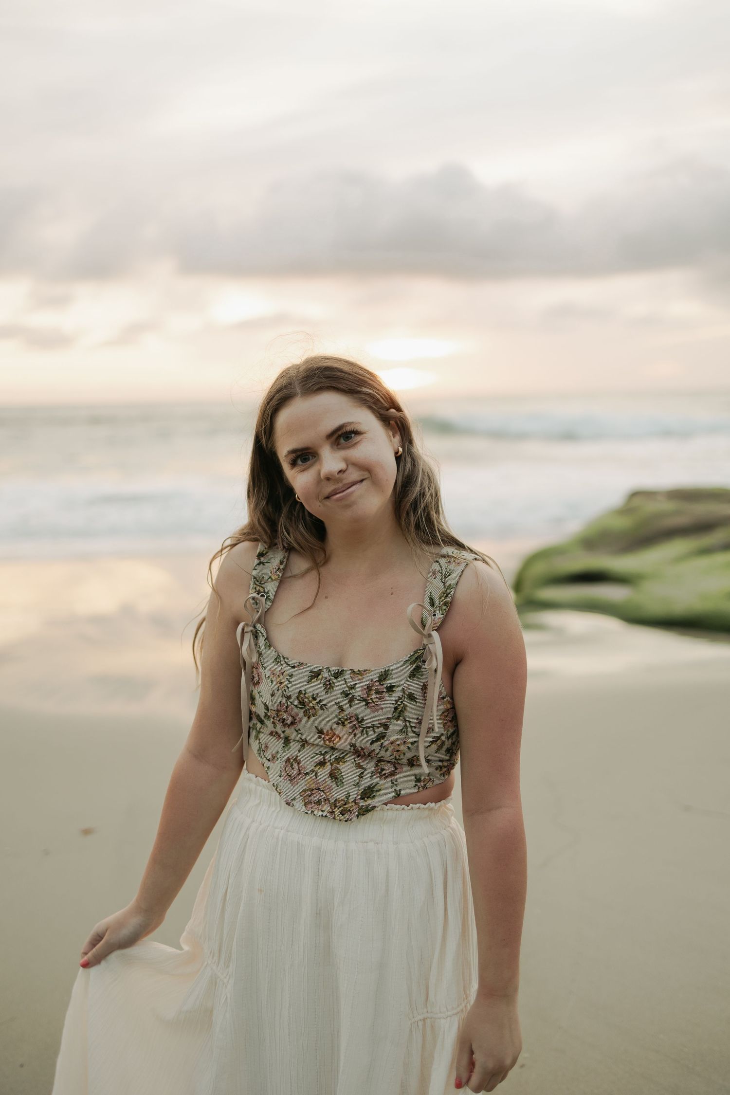 Person in a white floral dress poses on a misty beach at dusk.