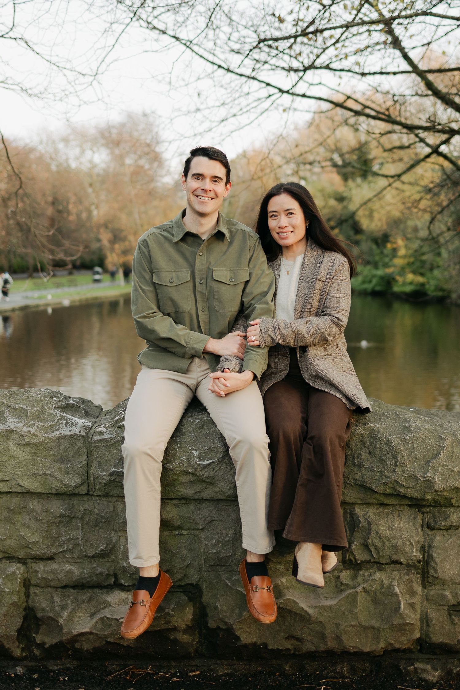 A smiling couple sits on a stone wall in an autumn park, dressed in smart casual outfits, with a serene pond behind them.