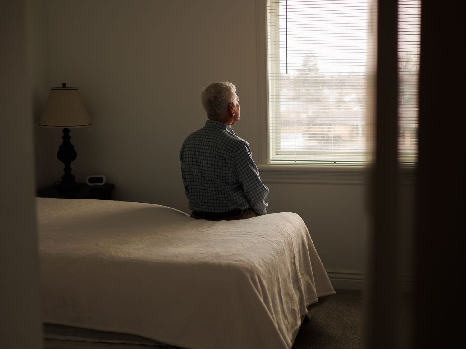Elderly man sitting alone on bed gazing out window in dimly lit bedroom, depicting loneliness and isolation.