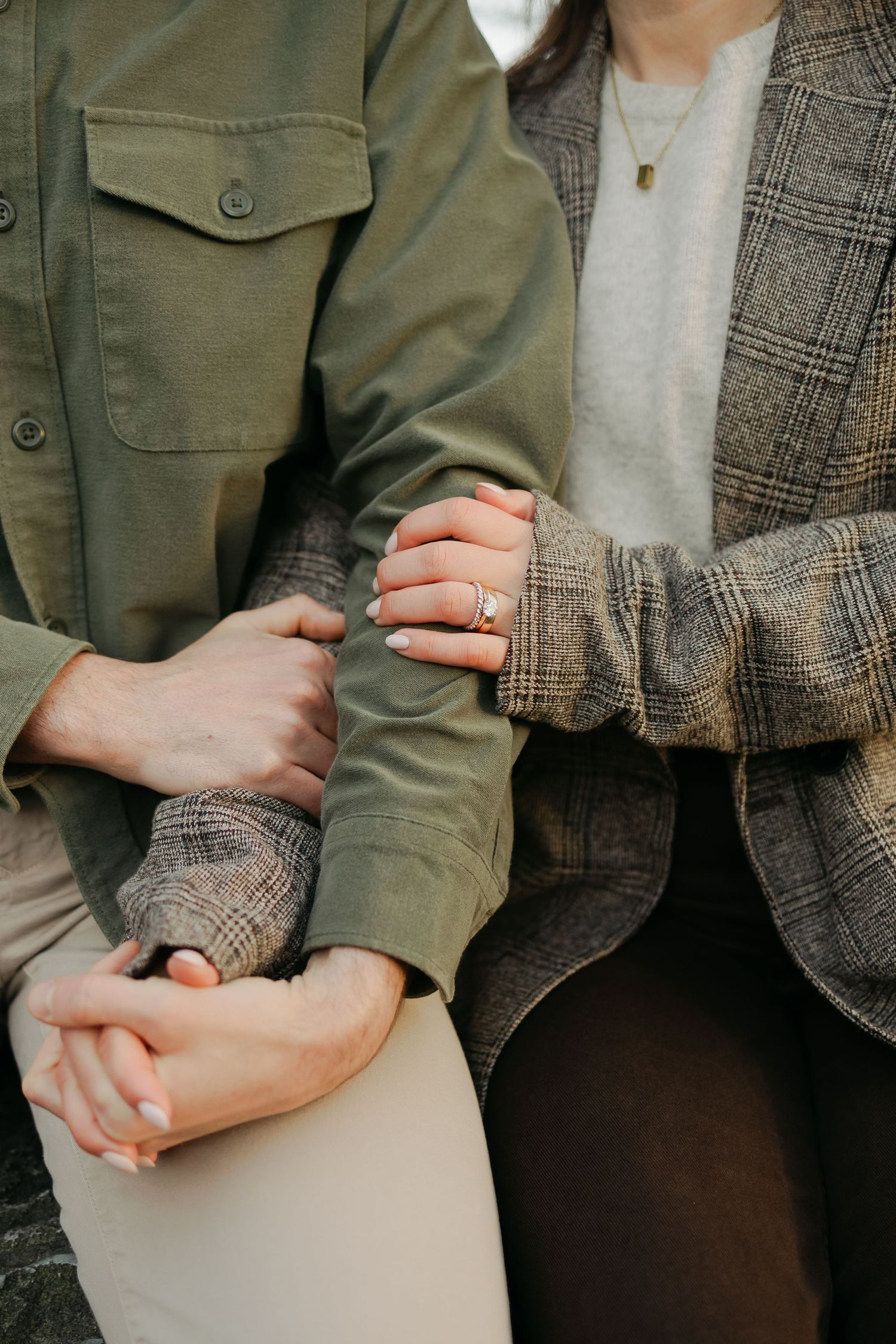 Close-up of couple holding hands, woman wearing engagement ring, dressed in green and plaid jackets.