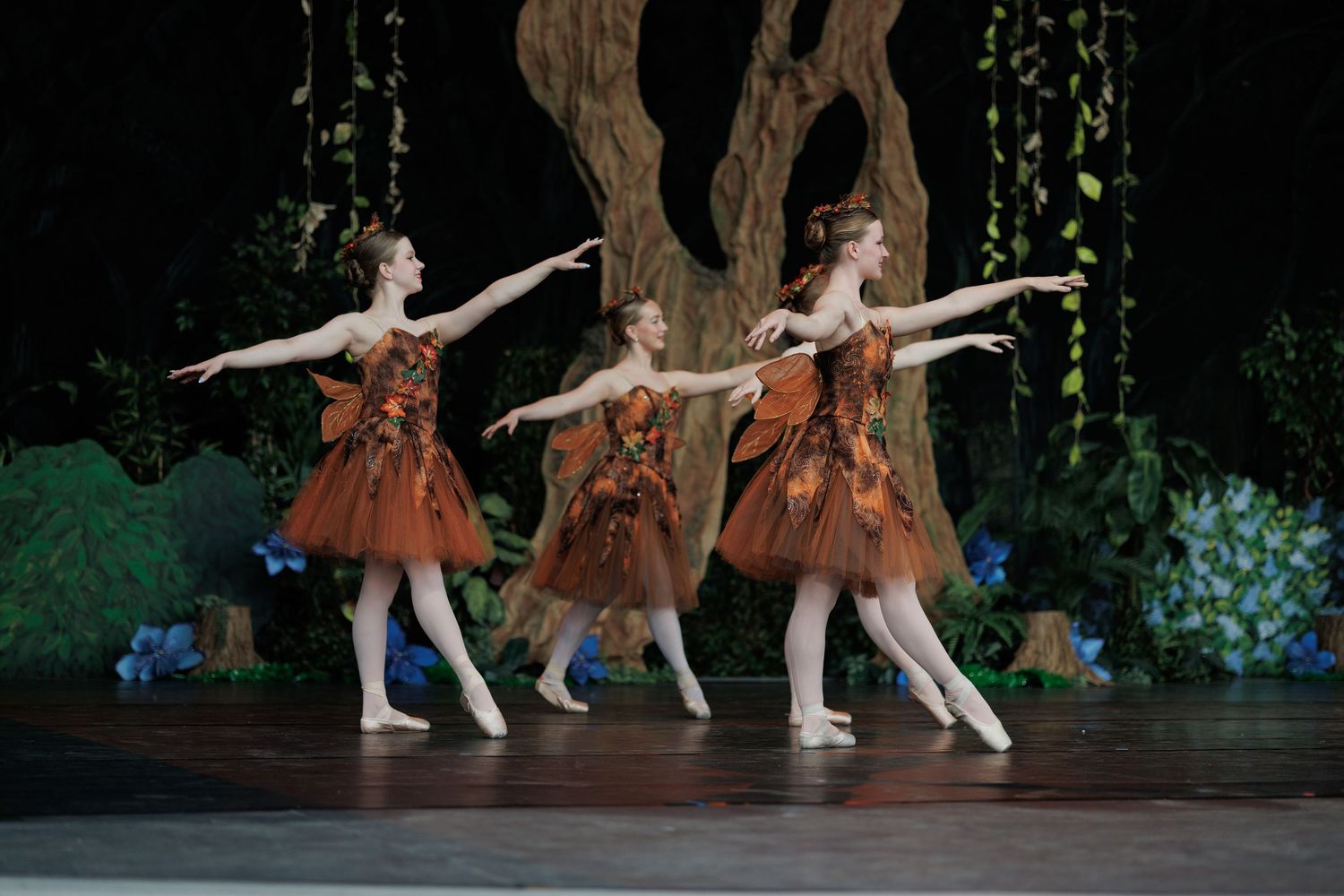 Three ballet dancers in brown costumes perform synchronized movements on stage with forest scenery backdrop.