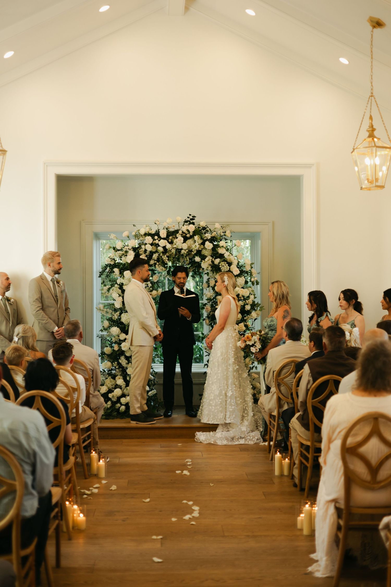 Wedding ceremony in progress inside bright white chapel with floral arch and seated guests.