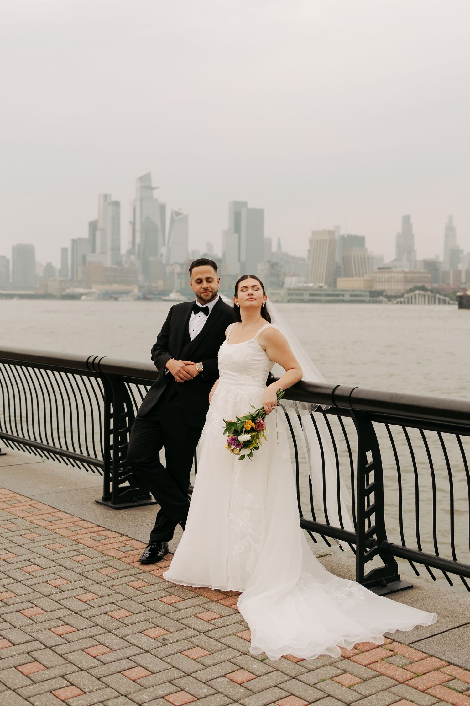 A couple poses on brick paving along an urban waterfront railing with dramatic skyline views.
