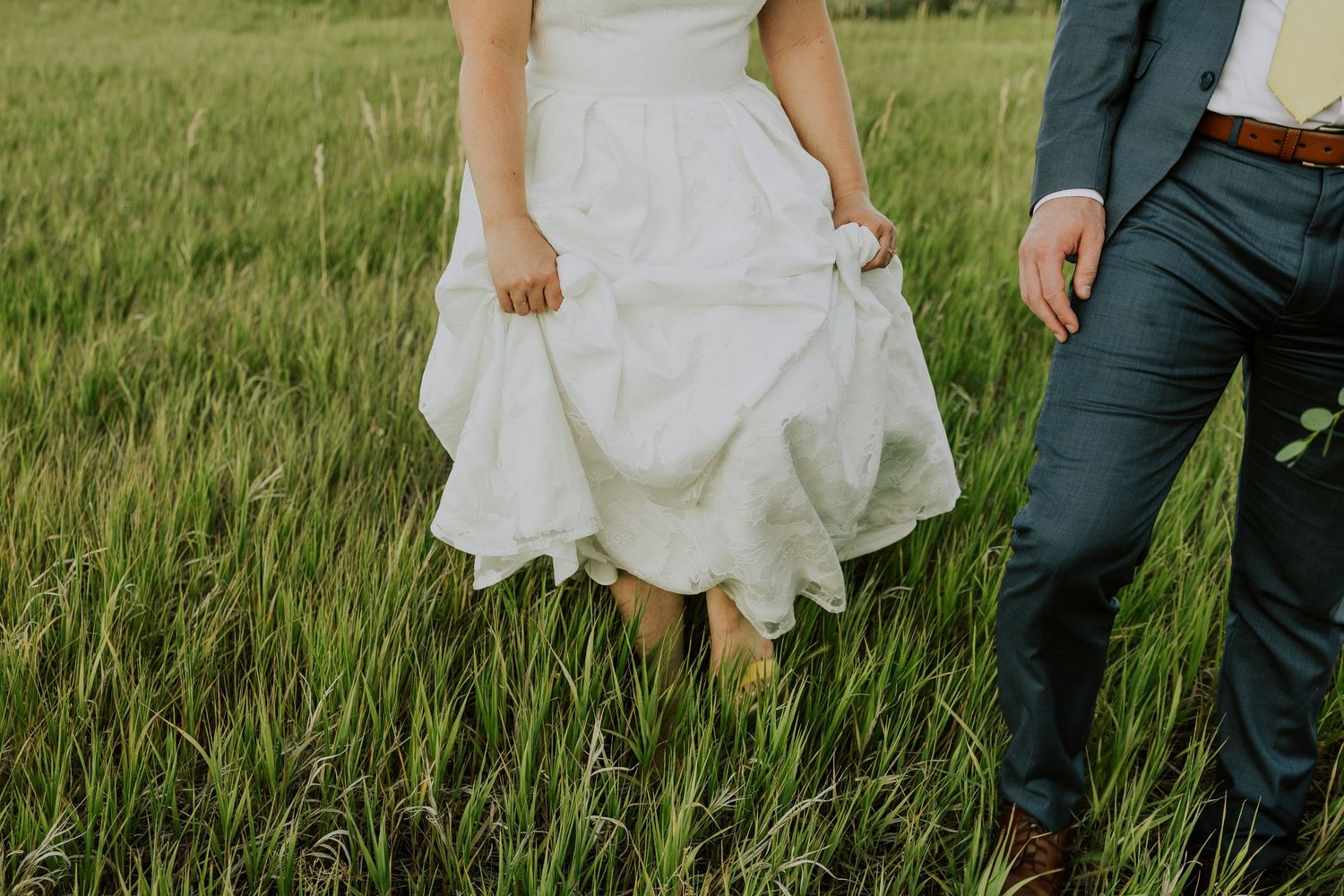 A white wedding dress and navy blue suit pants seen in a lush green meadow on a sunny day.