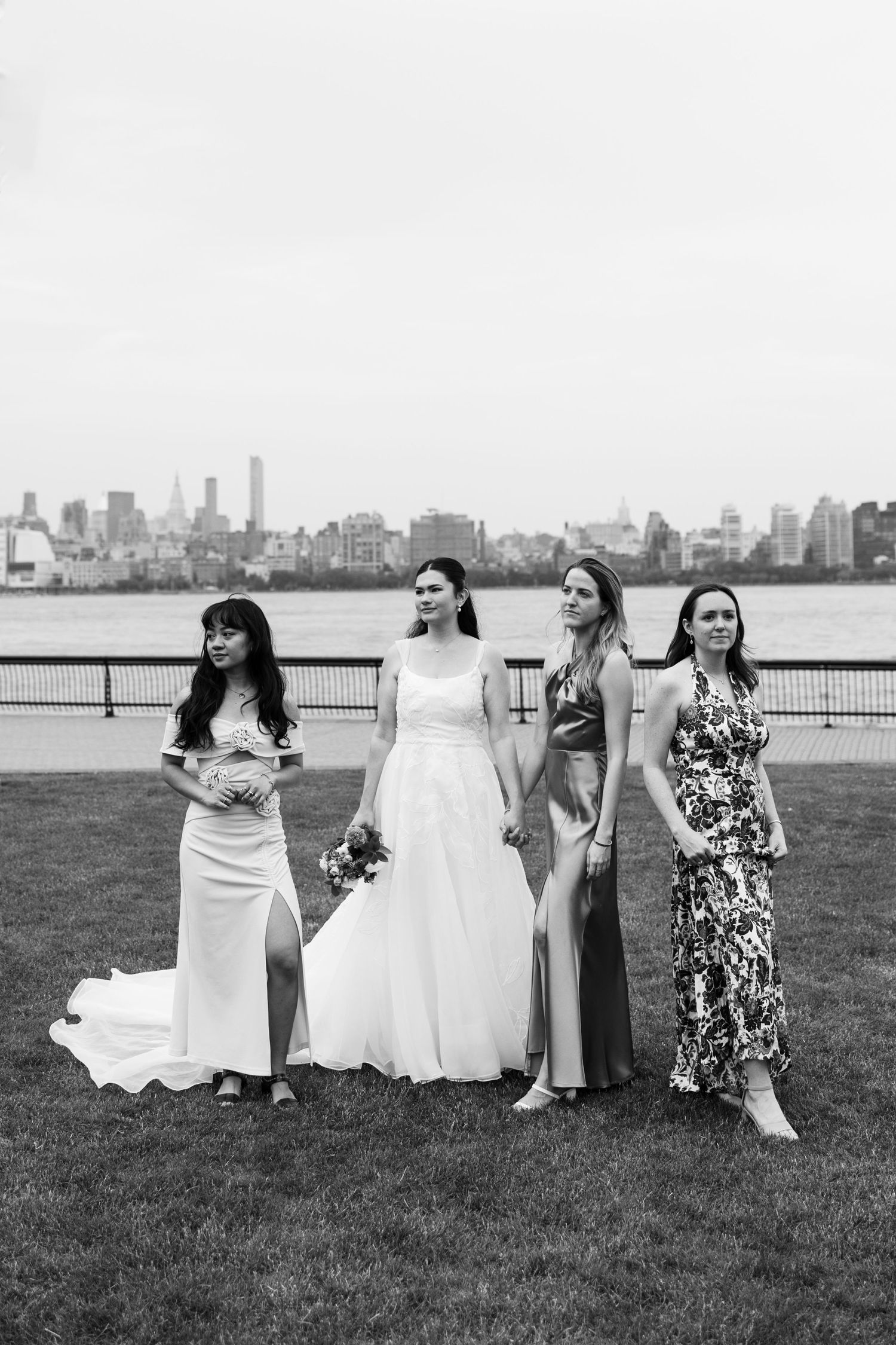 A black and white photo of a wedding party with bridesmaids and bride posing by the waterfront with city skyline.