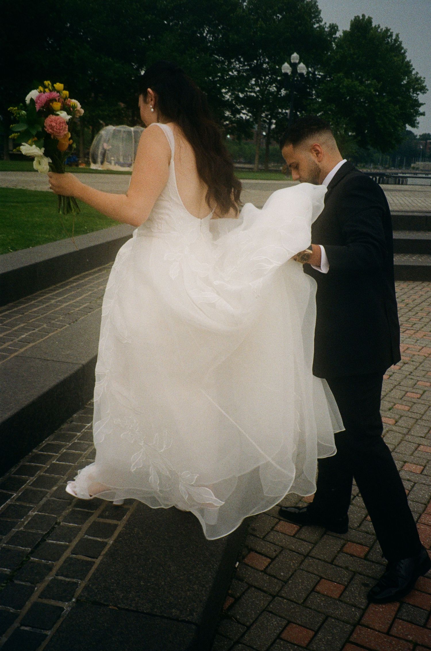 A bride in a flowing white dress holds a bouquet while walking with a groom in a black suit on brick pavement.