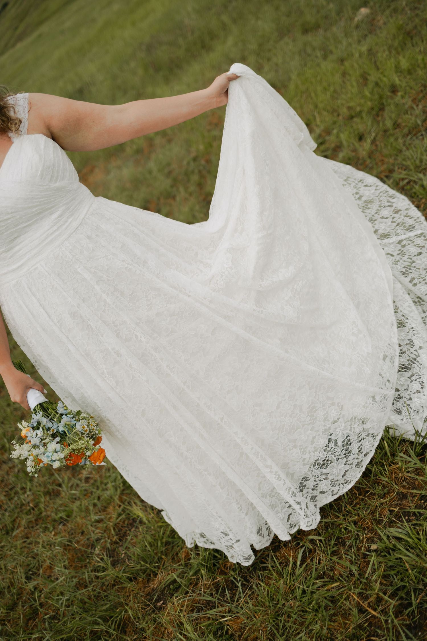 Bride in white lace wedding gown twirling on green hillside, holding bouquet of wildflowers outdoors.