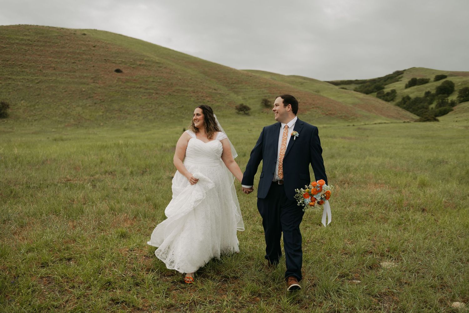 A bride and groom walk hand-in-hand through a lush green hillside meadow, she in white gown, he in navy suit.