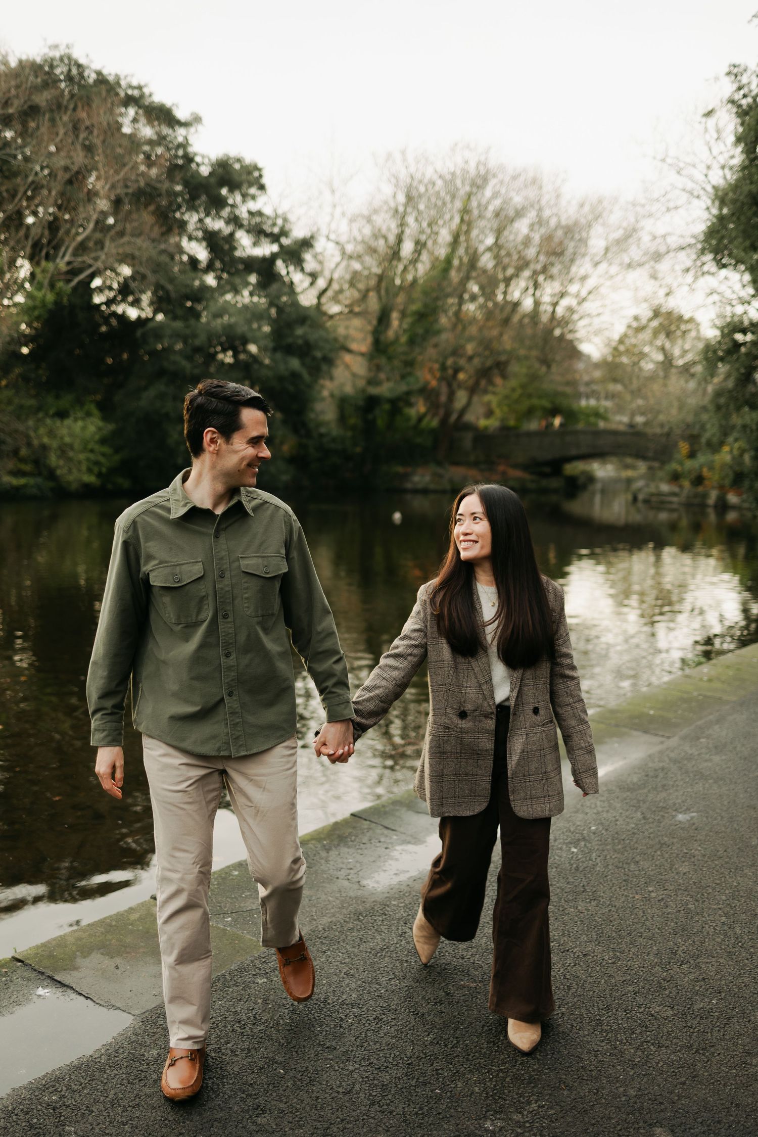 Couple holding hands walking along a scenic park pathway beside a calm river surrounded by autumn trees.