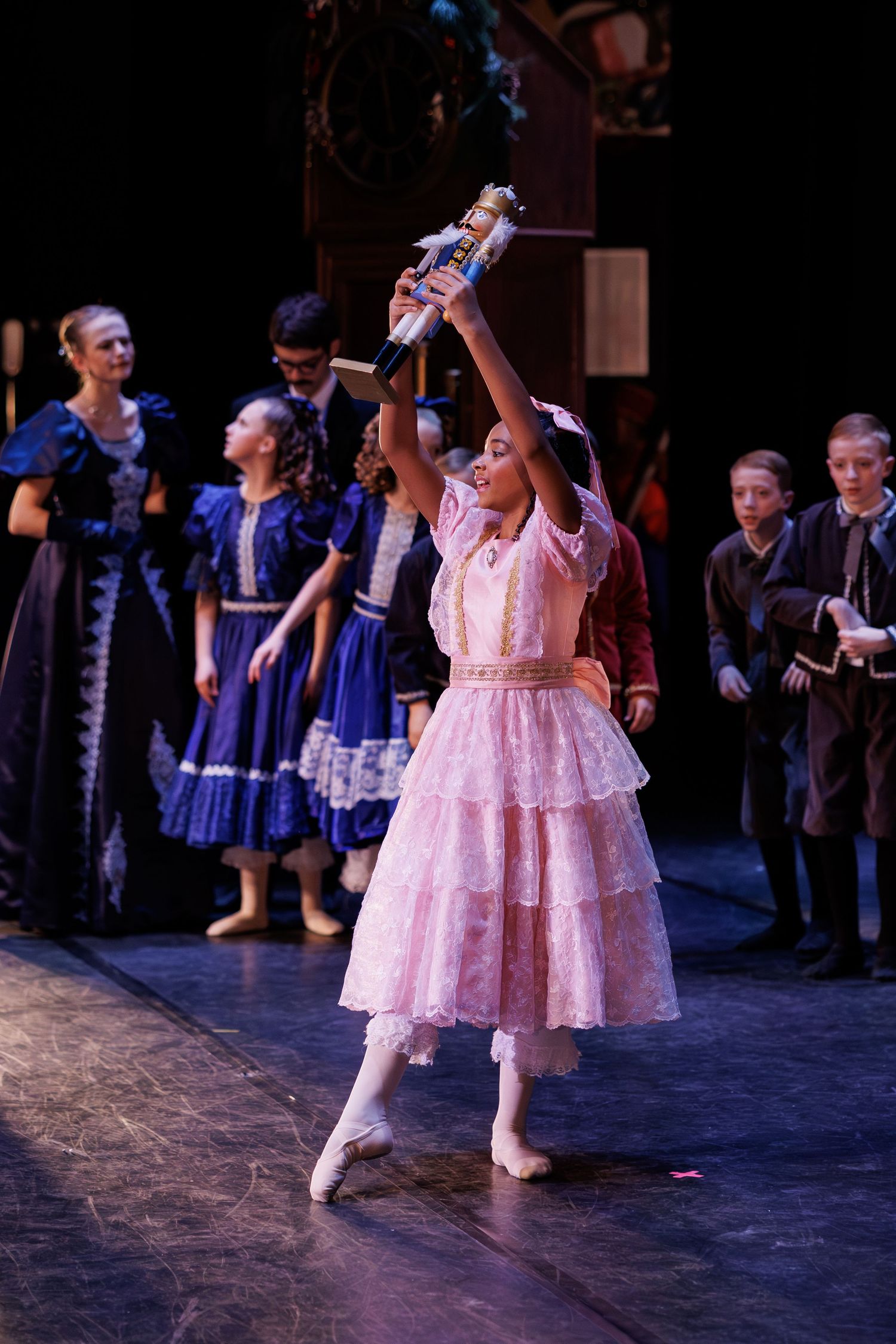 Ballerina in pink ruffled dress performs on stage during Nutcracker ballet with other performers in period costumes.