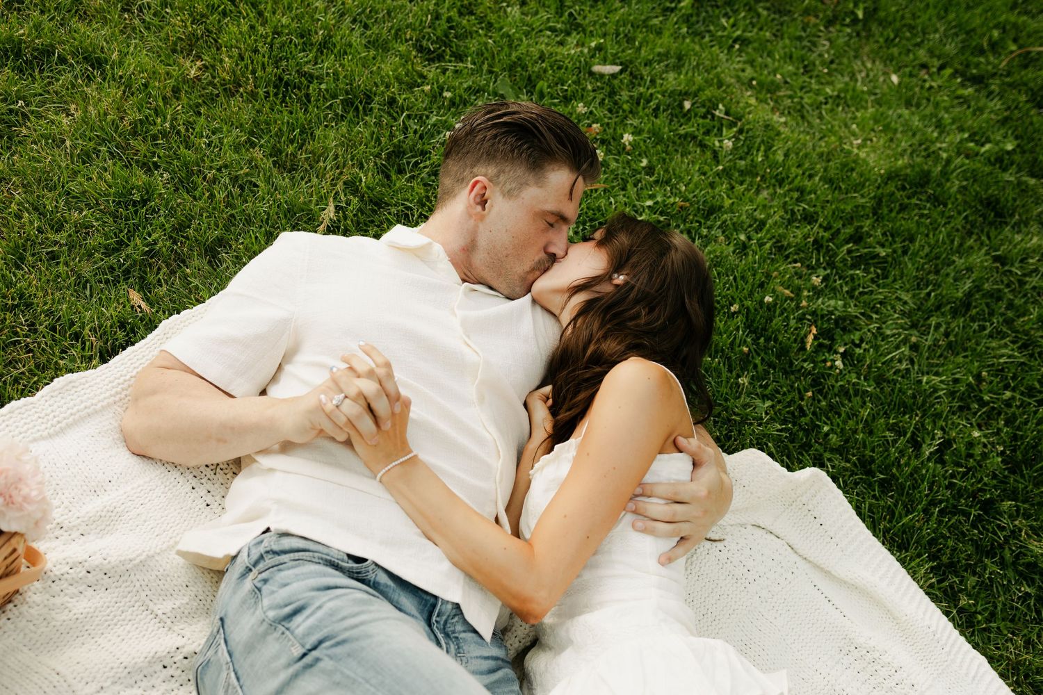 A romantic couple laying on a white blanket in green grass sharing an intimate moment.