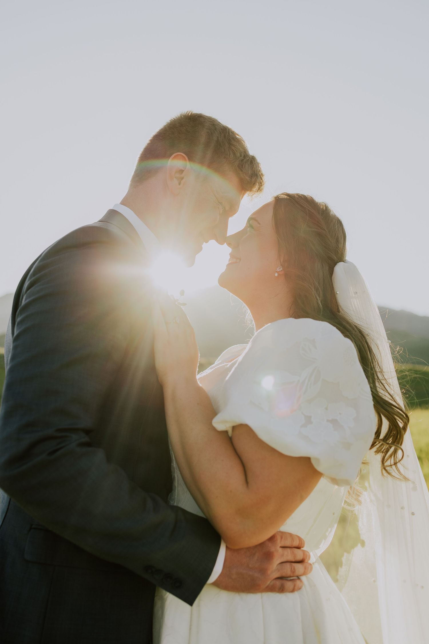 Silhouetted romantic couple shares a gentle kiss outdoors with bright sunlight creating a dreamy backlit glow.
