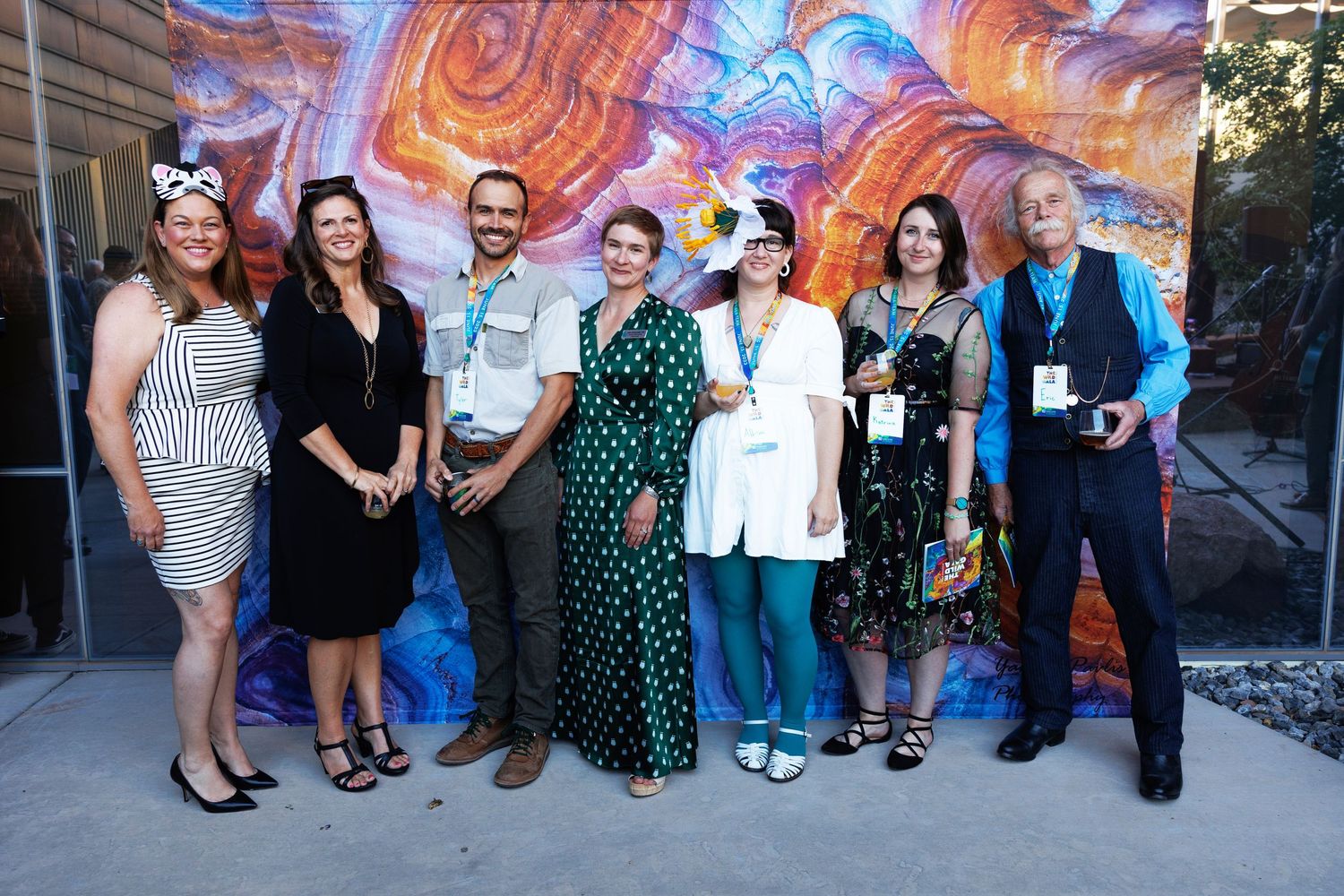 Group of seven people posing together outdoors in front of a colorful mural at an event.