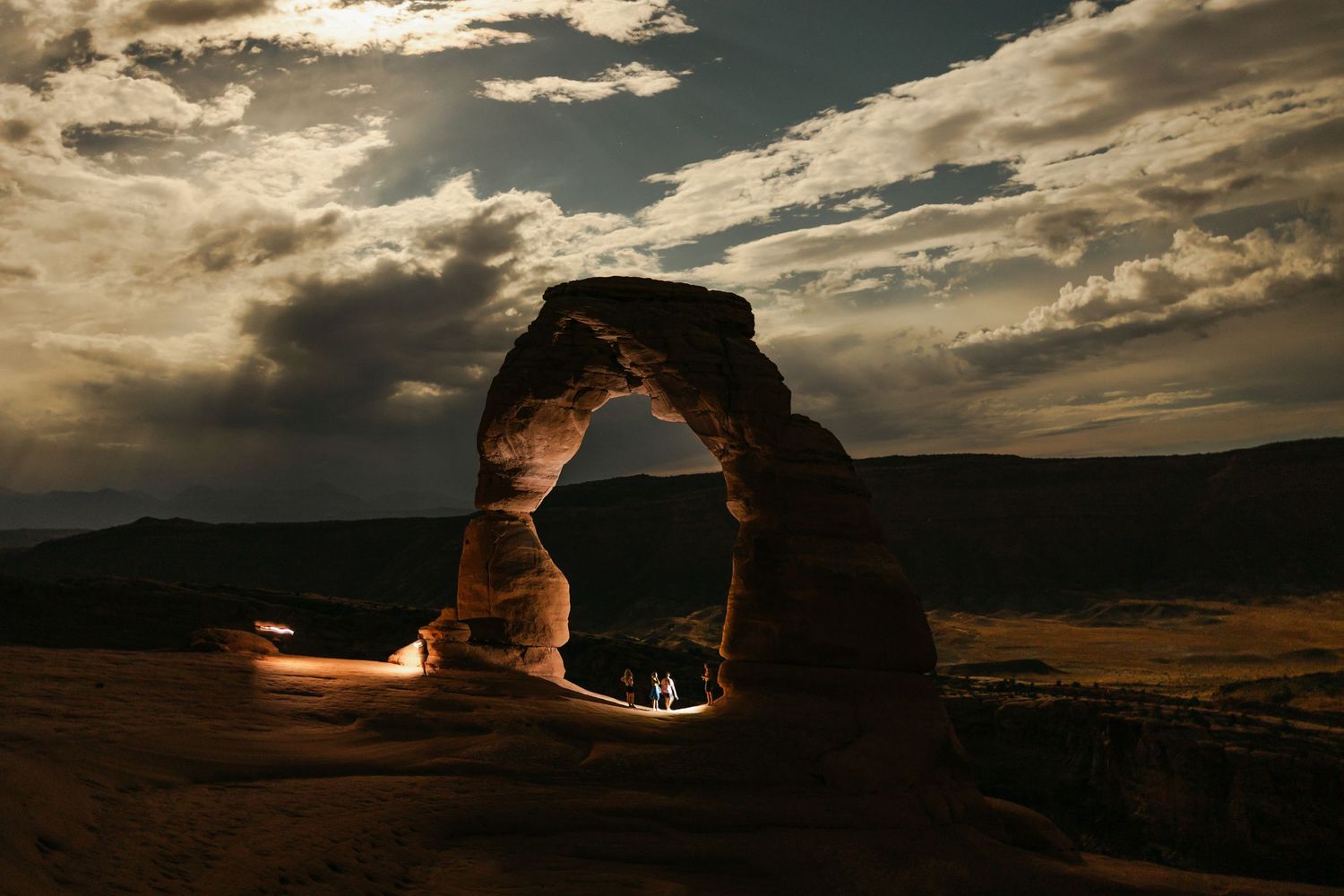 Dramatic silhouette of Delicate Arch at dusk with glowing light beneath and stormy clouds over Utah desert landscape.