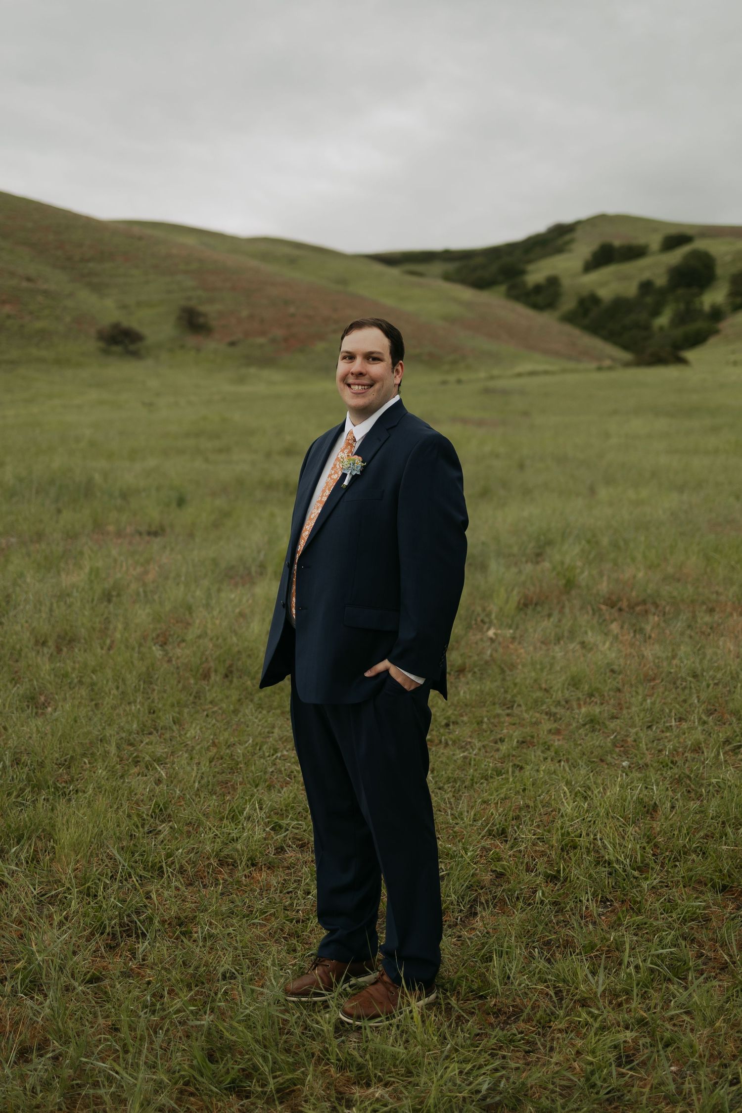 Smiling groom in navy suit and floral tie stands in lush green rolling hills on his wedding day.