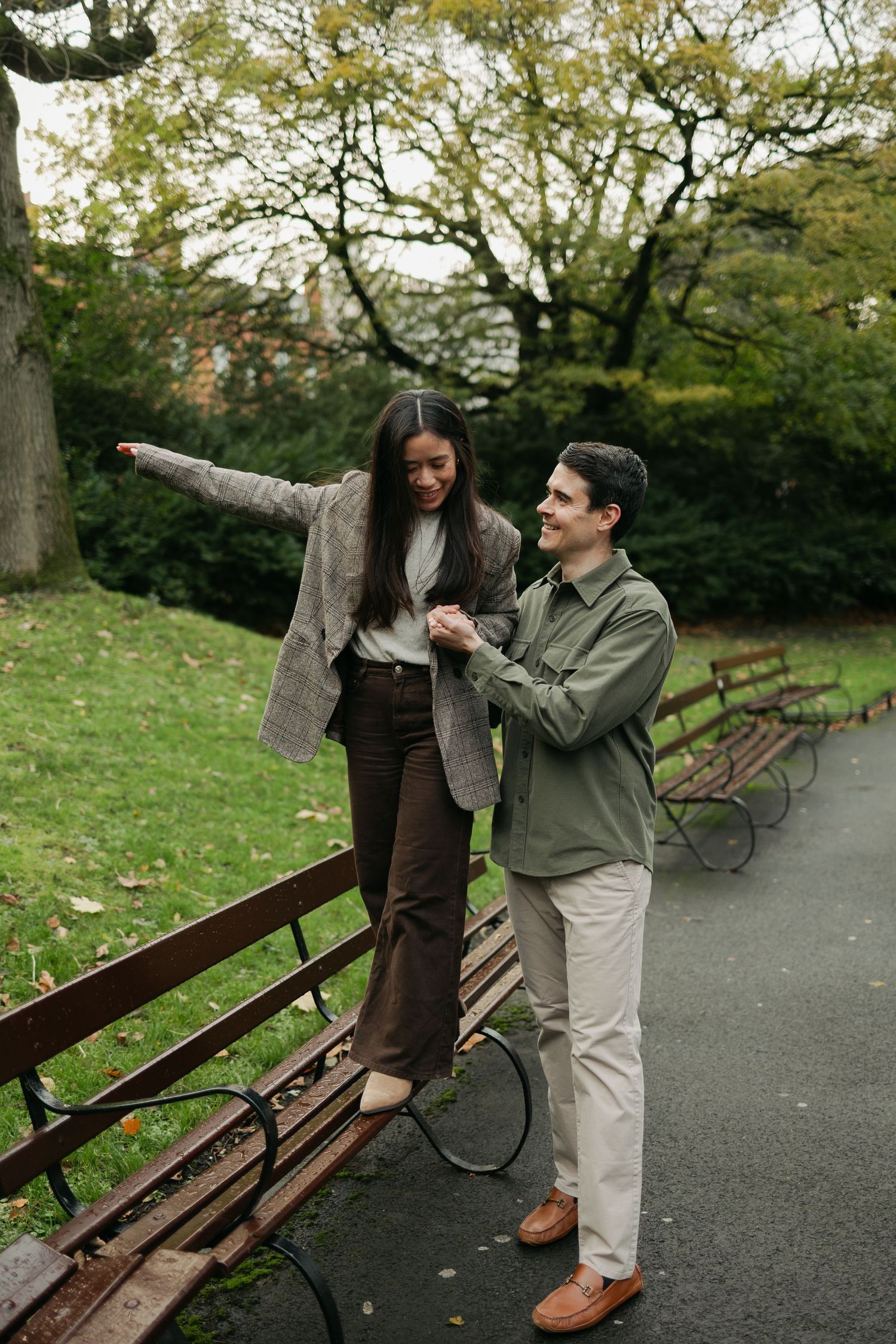 A smiling couple in a park, woman balancing on a bench holding mans hand, wearing autumn outfits.