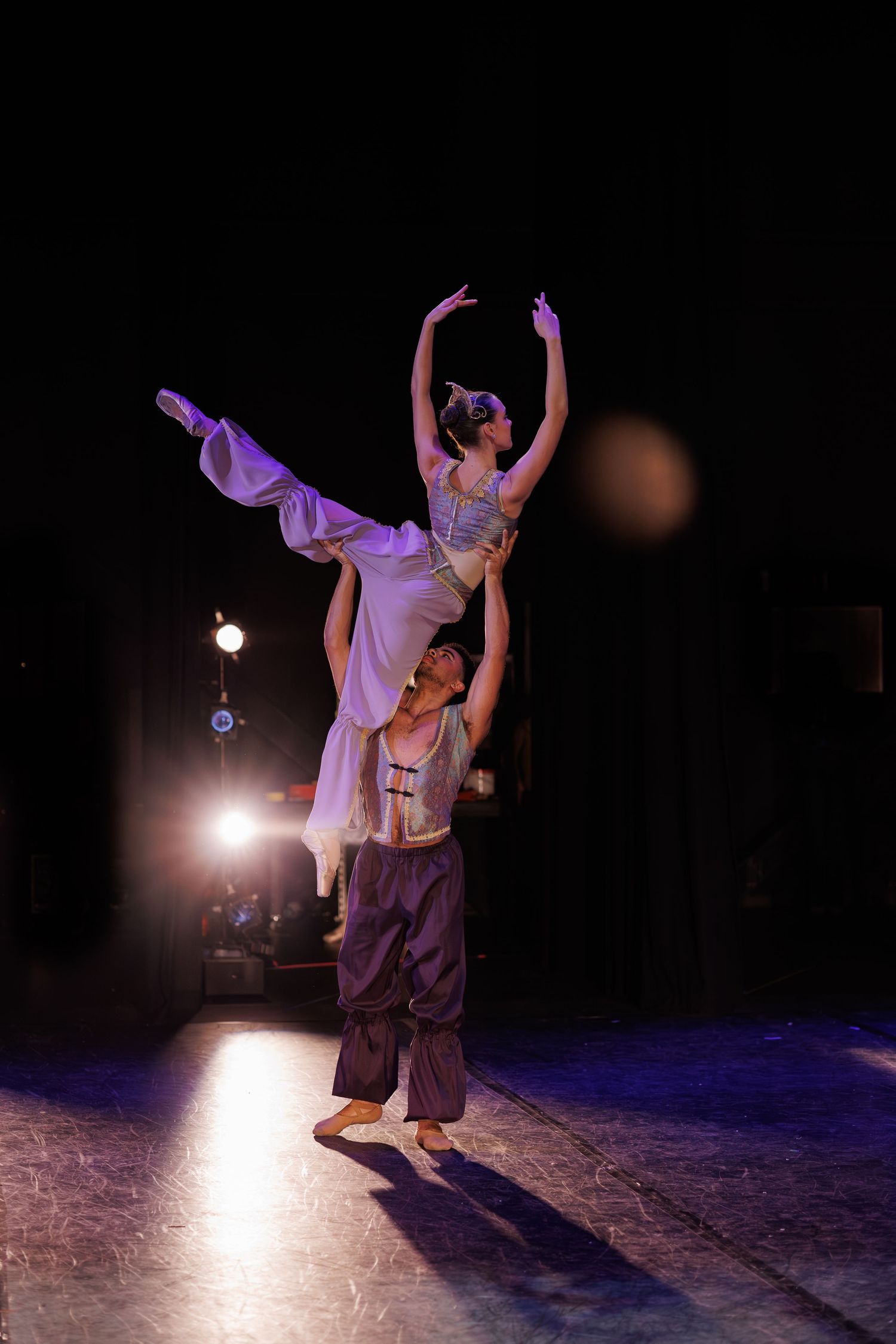 Dancers in purple and white outfits perform expressive movements under dramatic stage lighting.