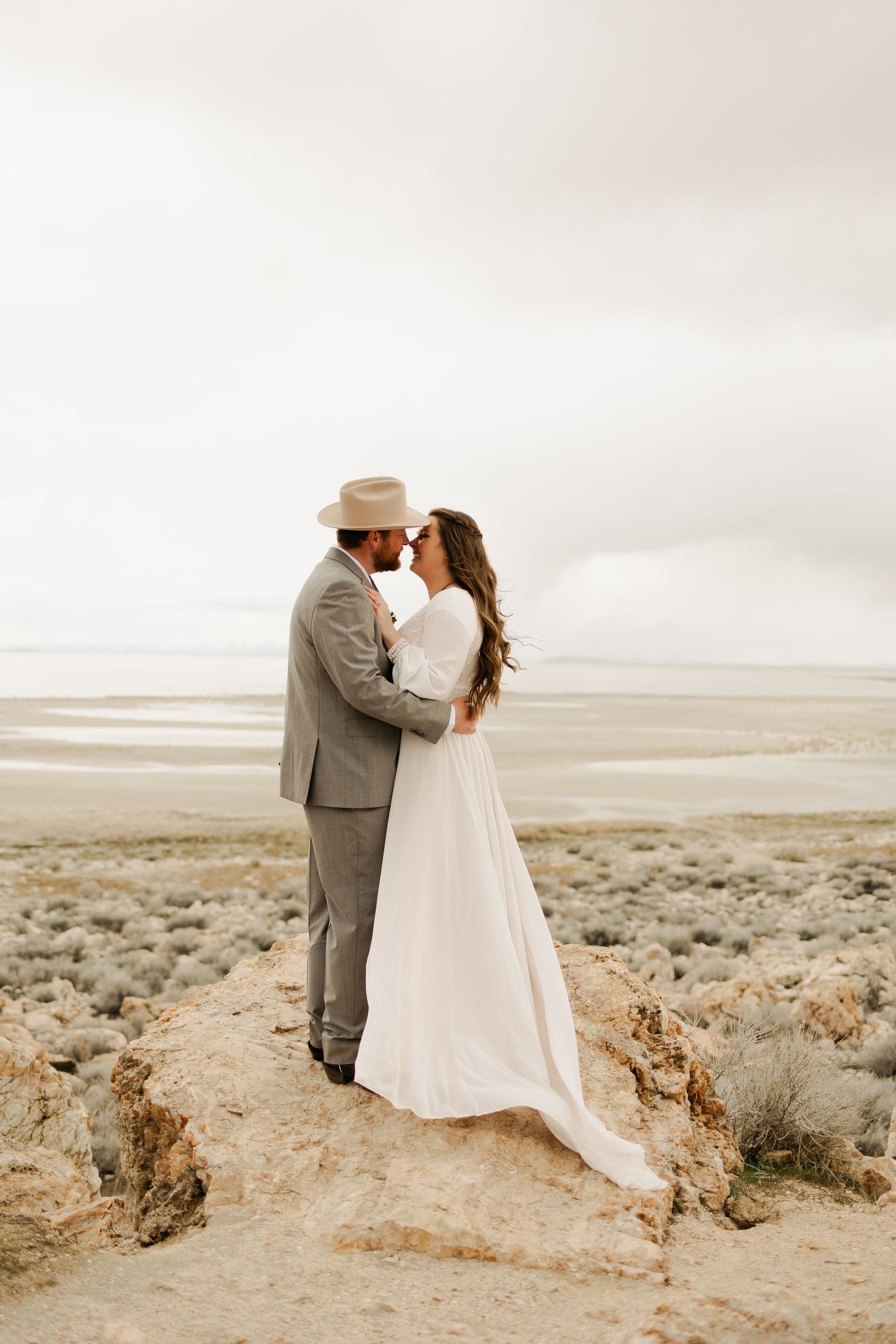 Newlyweds embrace against a desert landscape with flowing white dress and grey suit.