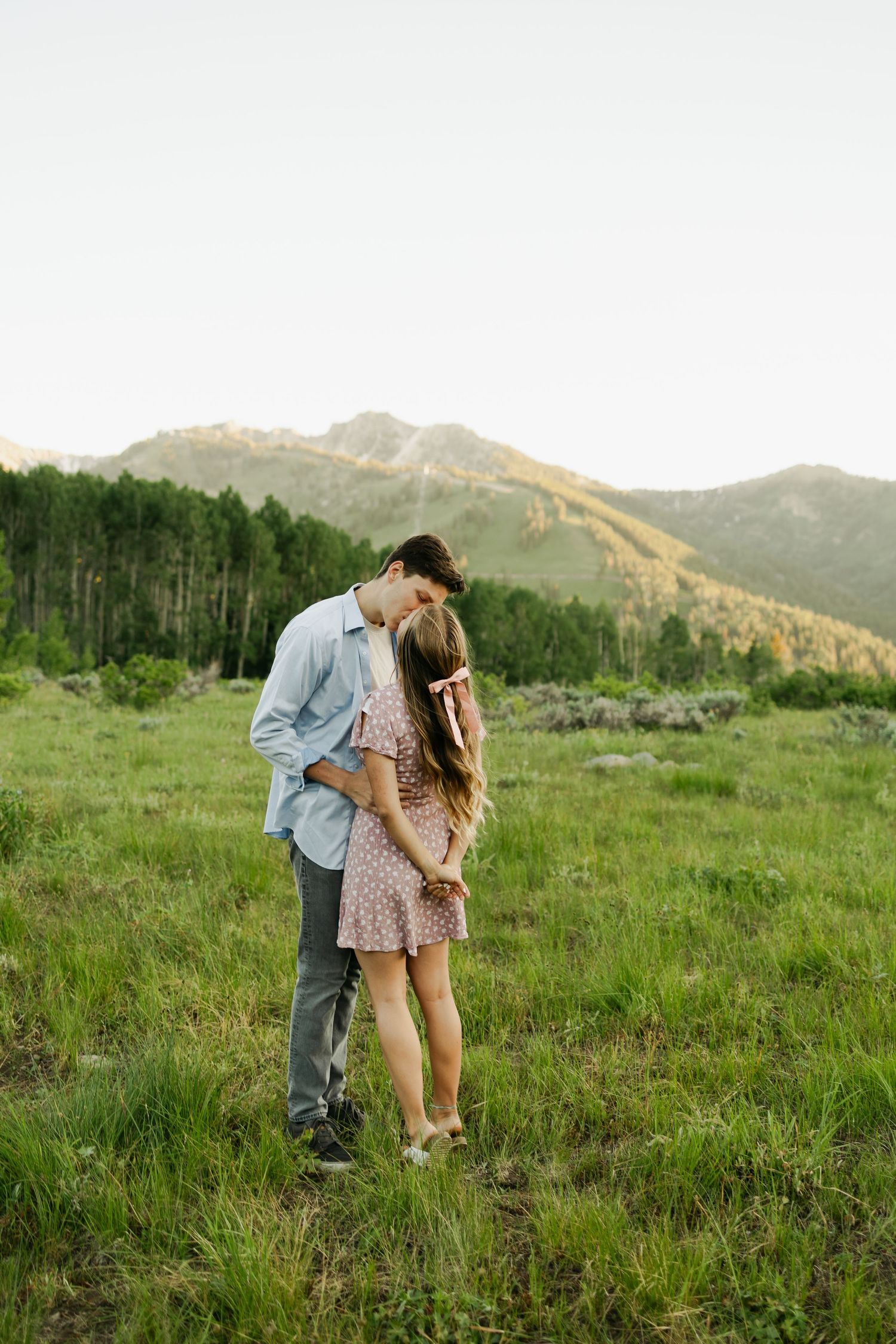 Couple sharing tender moments in mountain meadow with scenic backdrop.