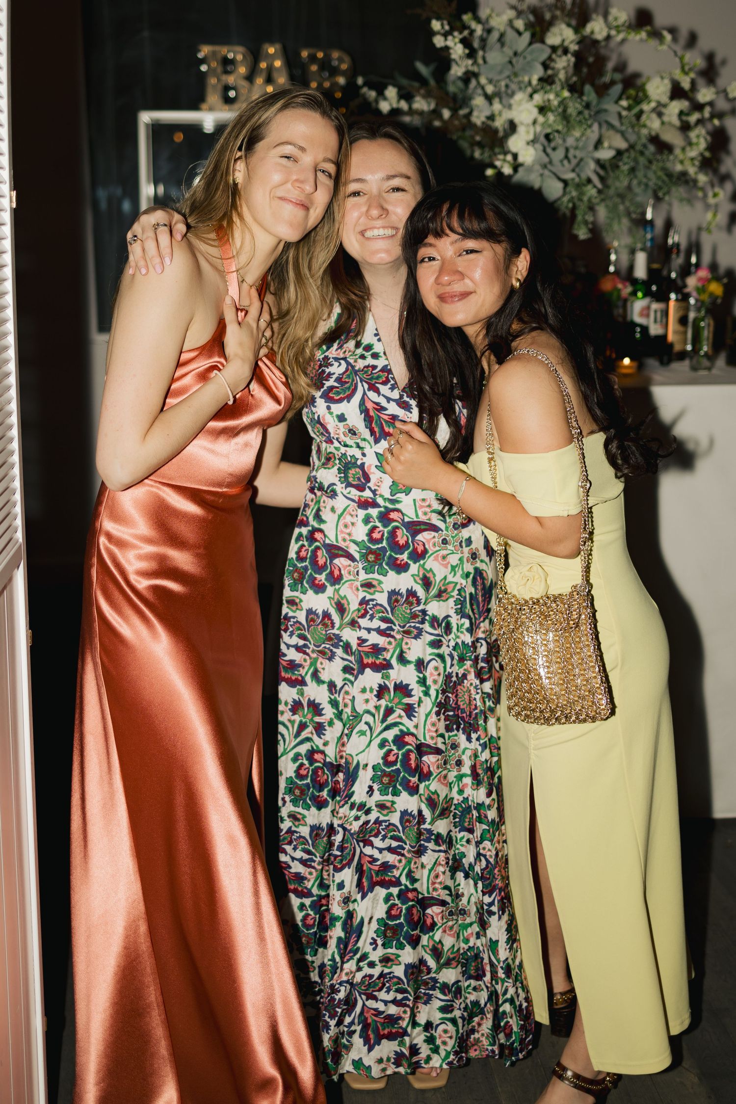 Three friends in elegant dresses pose together at evening wedding reception.