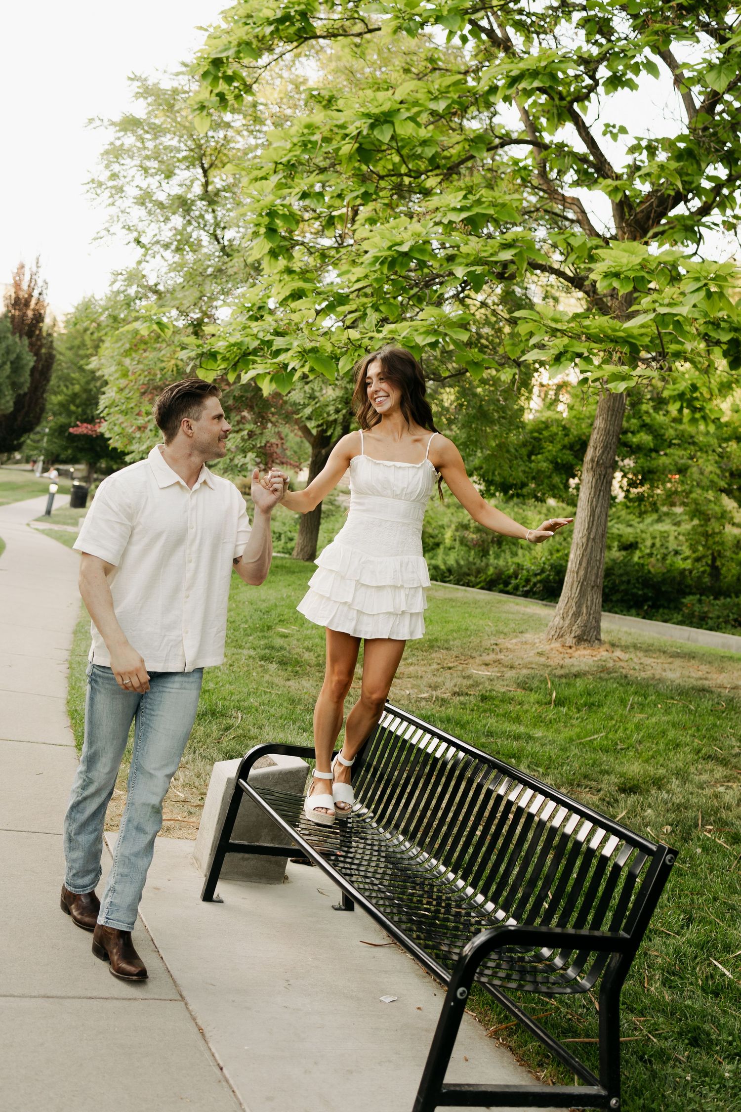 A couple shares a playful moment in a park as one person balances on a black metal bench while holding hands.
