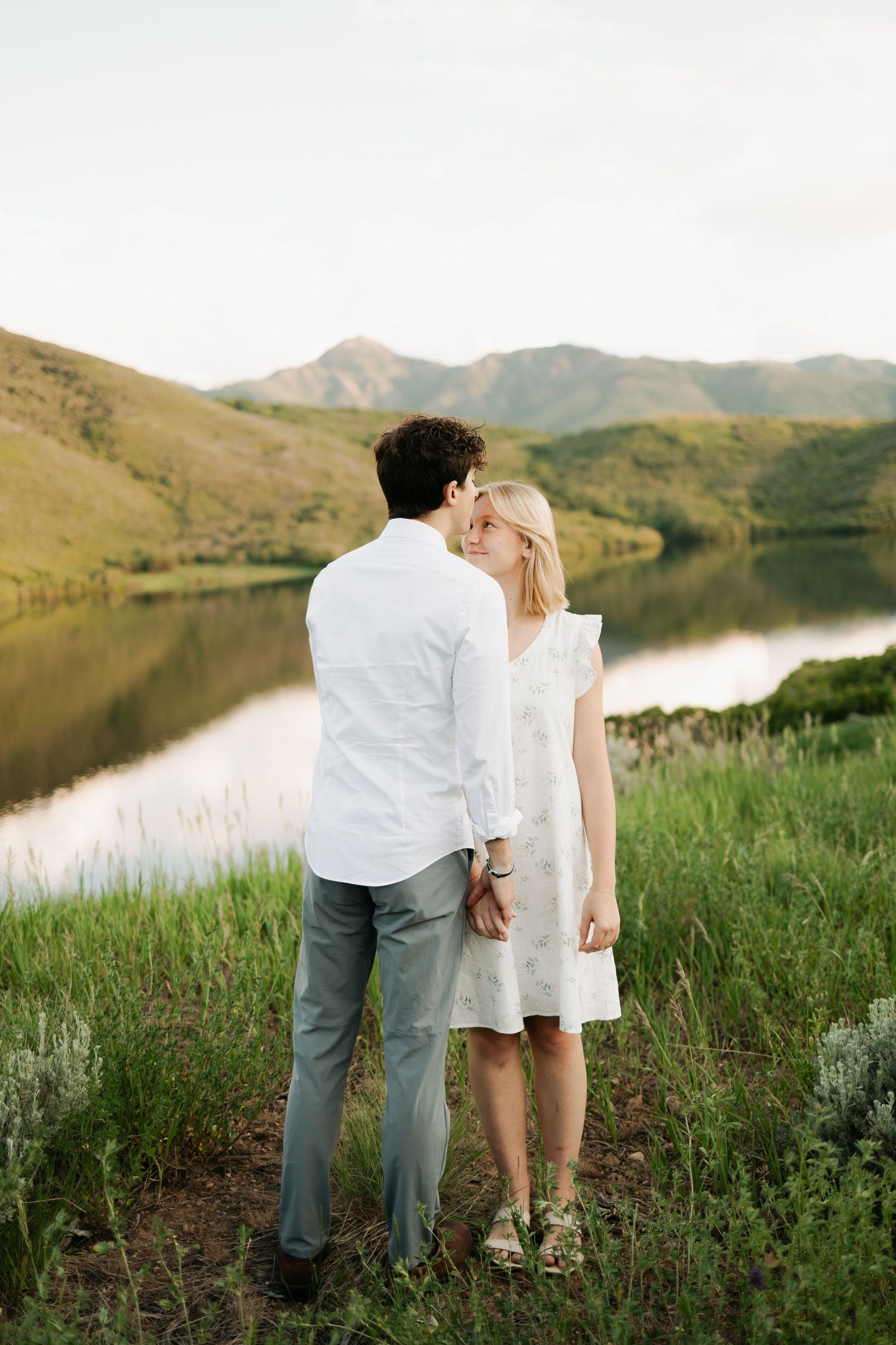A couple in light colored outfits stands by a serene mountain lake at sunset.