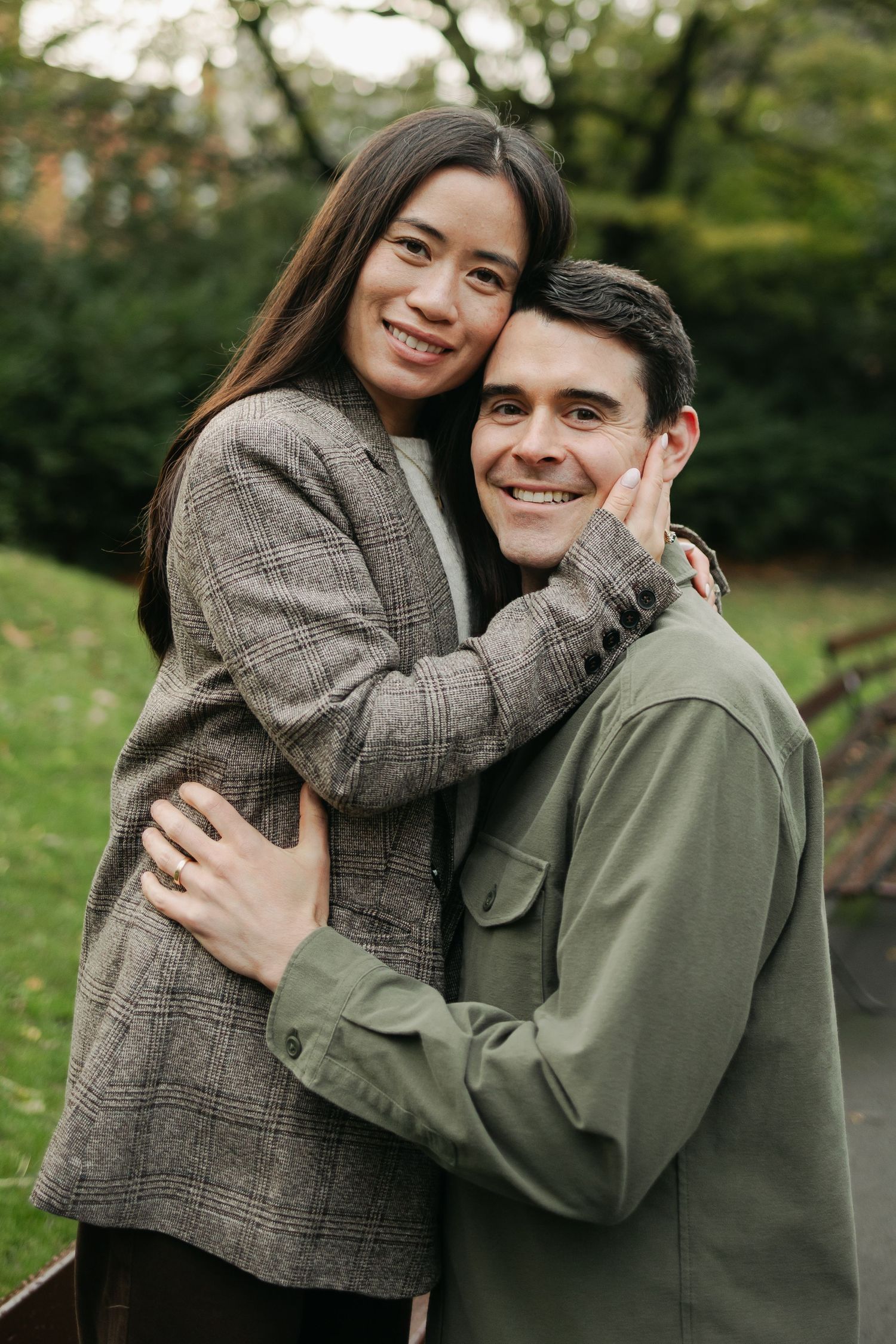 Happy interracial couple embracing in a park, woman in plaid blazer touching mans face while he smiles at camera.