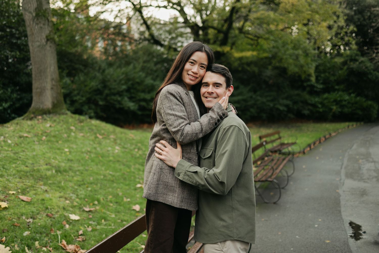 Couple embracing and smiling in a lush green park on an autumn day, surrounded by trees and a winding pathway.