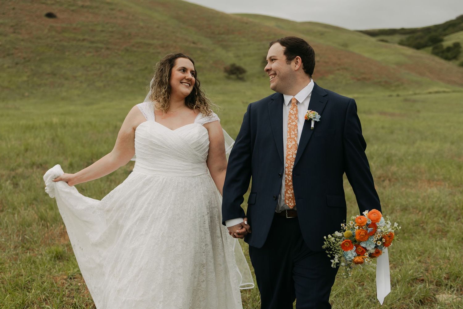 Bride and groom walk hand-in-hand through green hills, she in white gown, he in navy suit with orange floral tie.