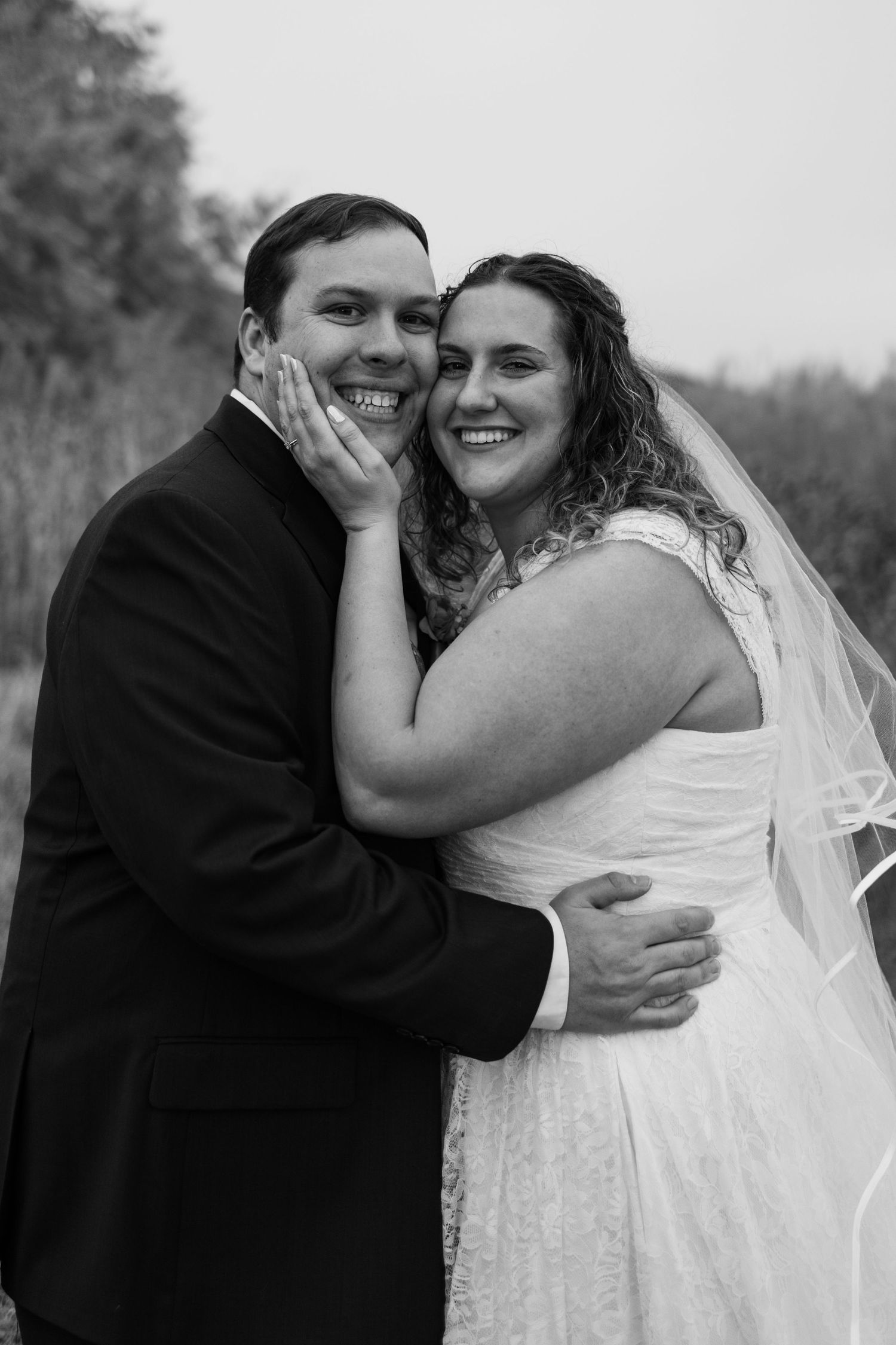 Black and white wedding portrait of smiling bride and groom embracing outdoors, bride wearing veil and lace gown.