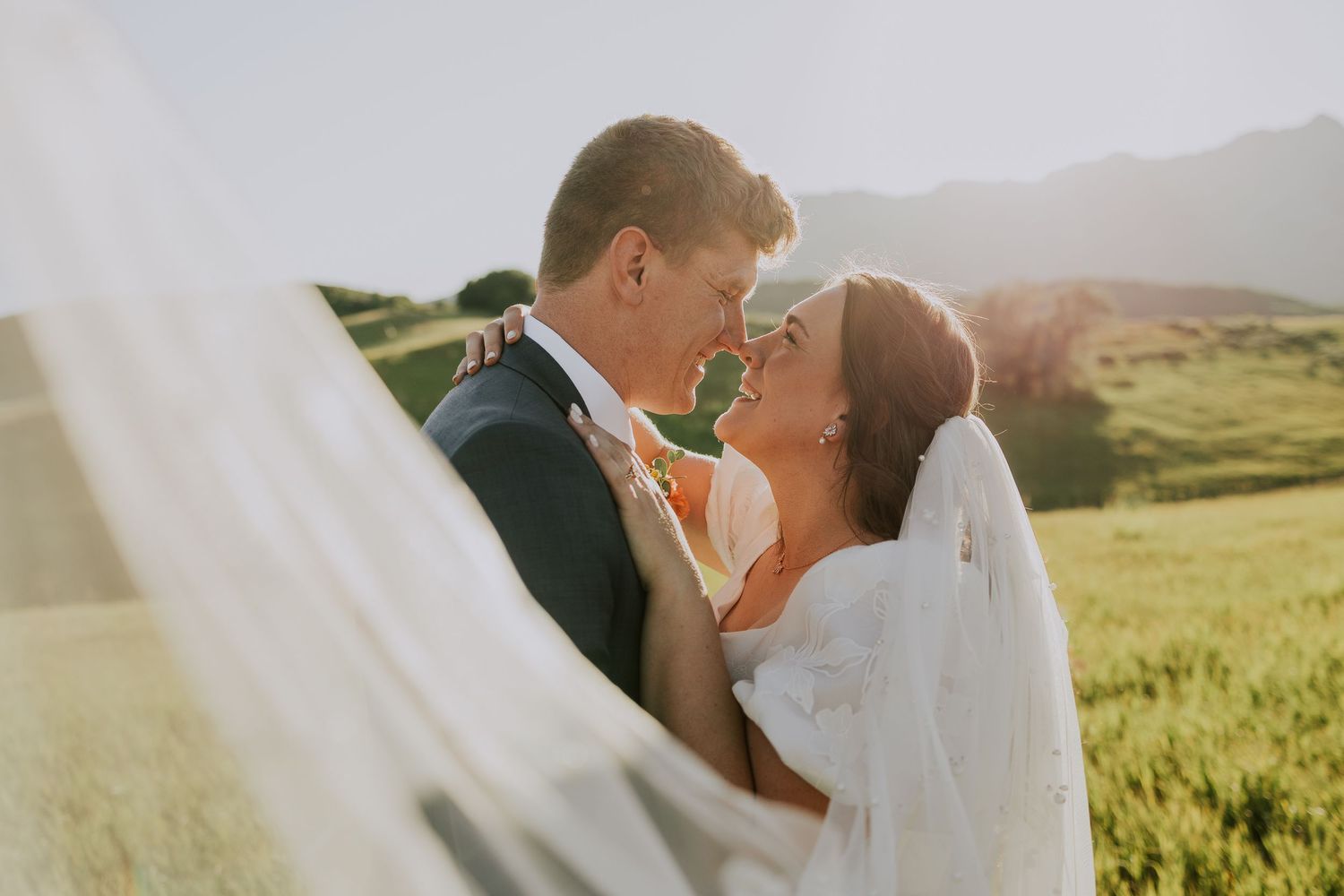 Newlywed couple embraces in sunlit meadow with flowing white veil and rolling hills in background.